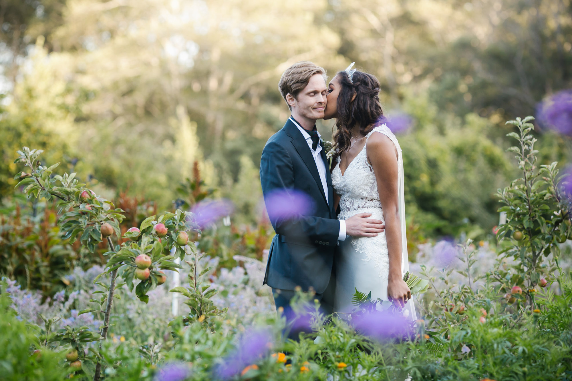 Bride in crystal crowns kisses her husband surrounded by flowers and apples
