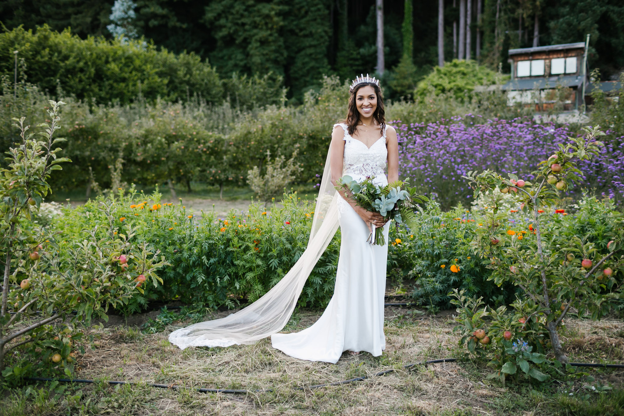 Bride in crystal crown and cape veil stands in flower field and orchard