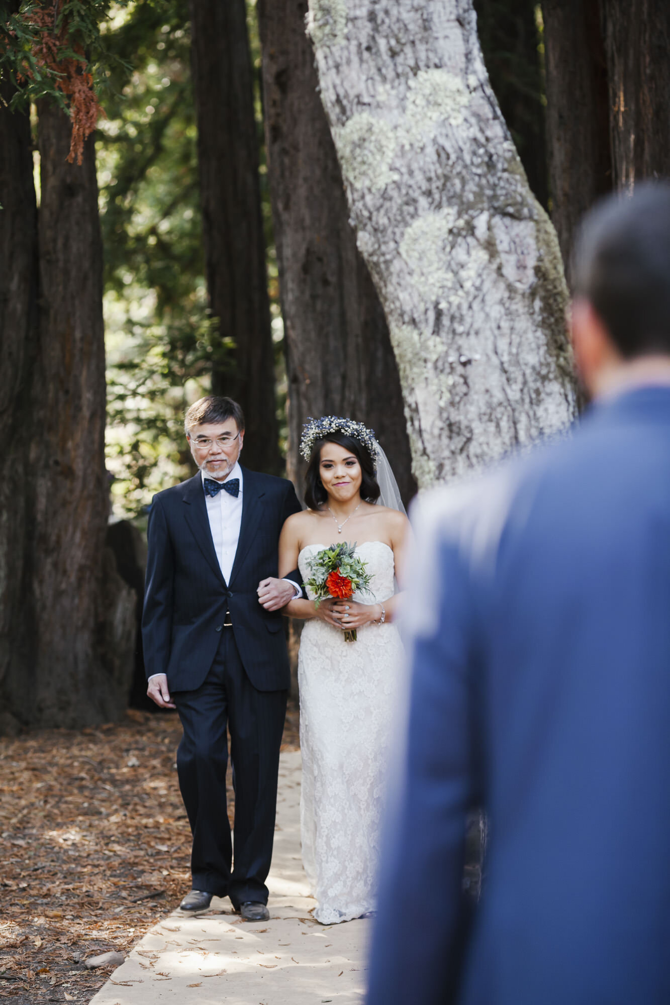 Smiling bride and her dad walk down the aisle in a redwood grove