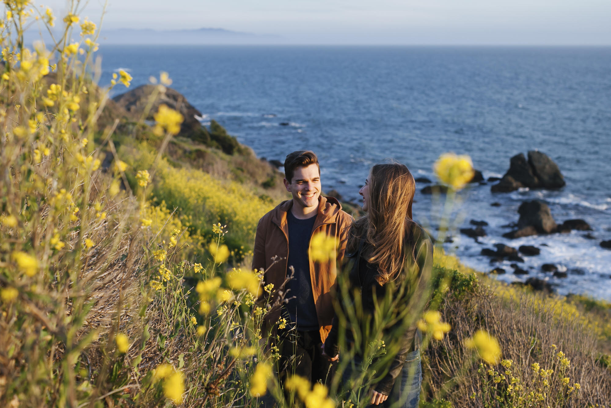 Engaged couple walk among yellow wildflowers along the California coast