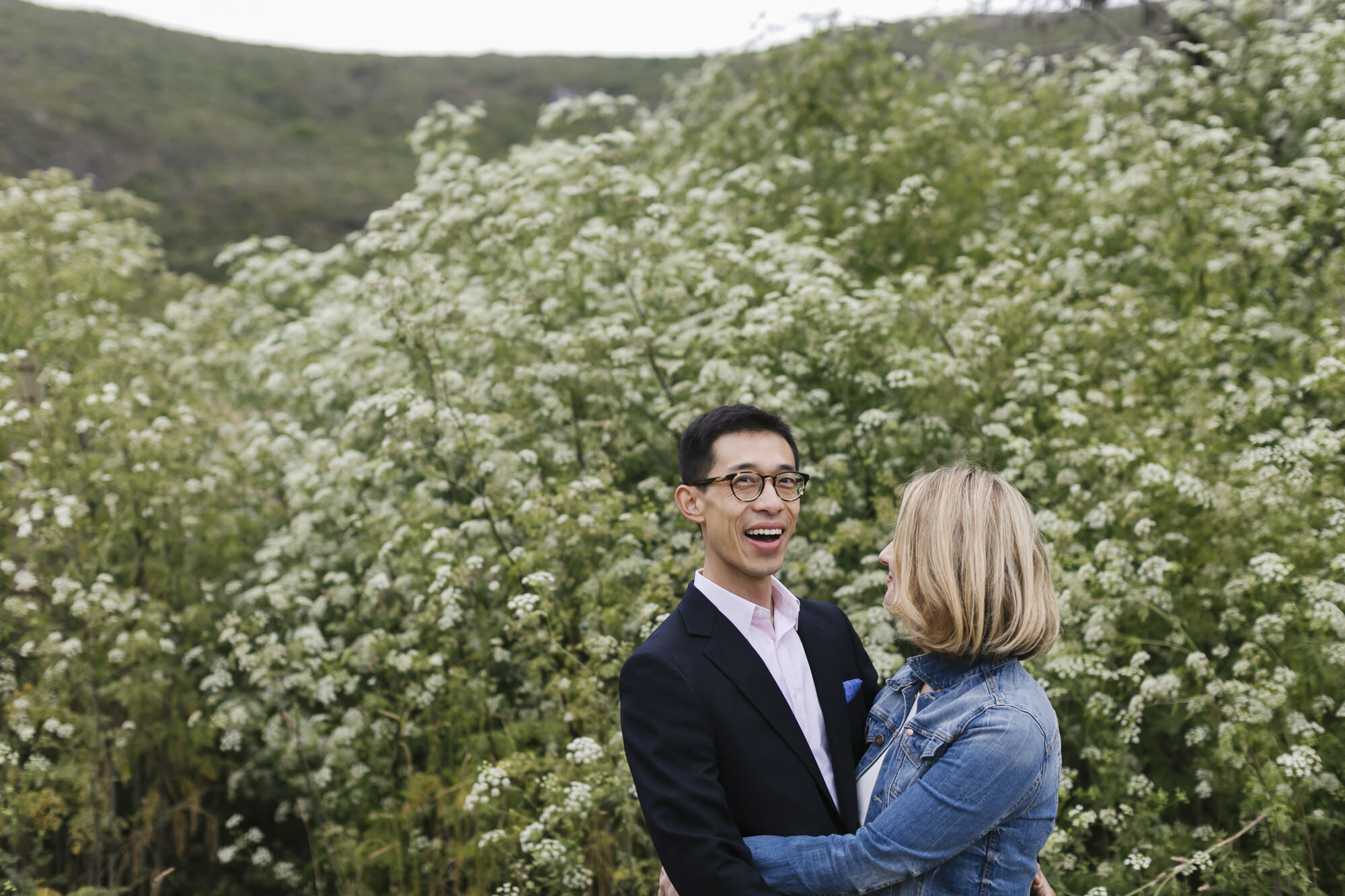 Groom smiles at the camera while hugging his wife with white wildflowers in the background