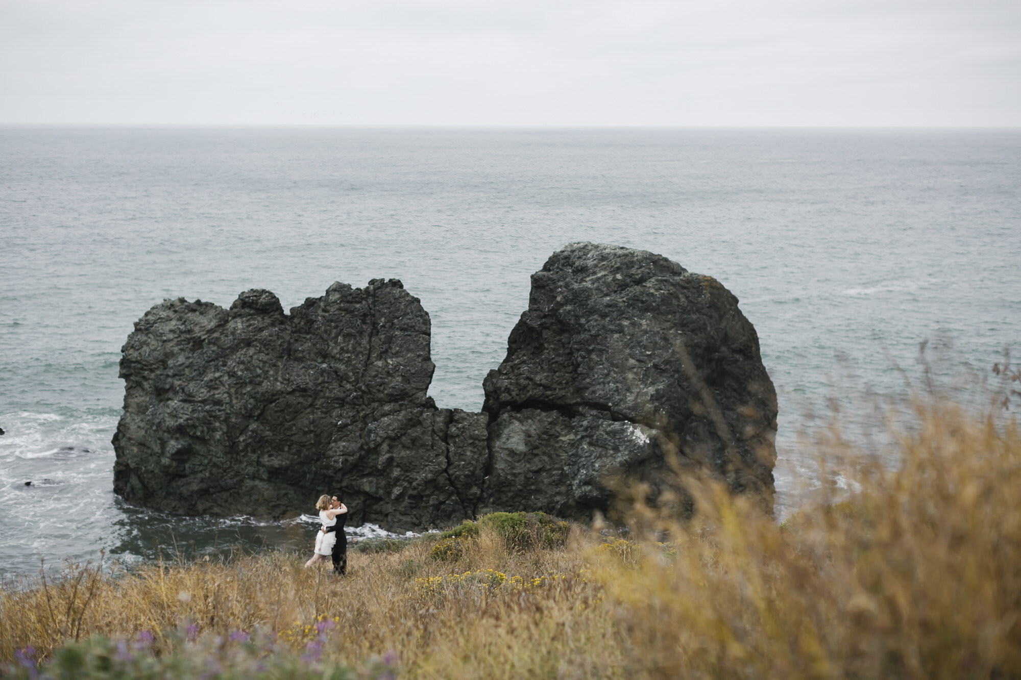 Married couple kiss in front of giant rock in the ocean along the California coast
