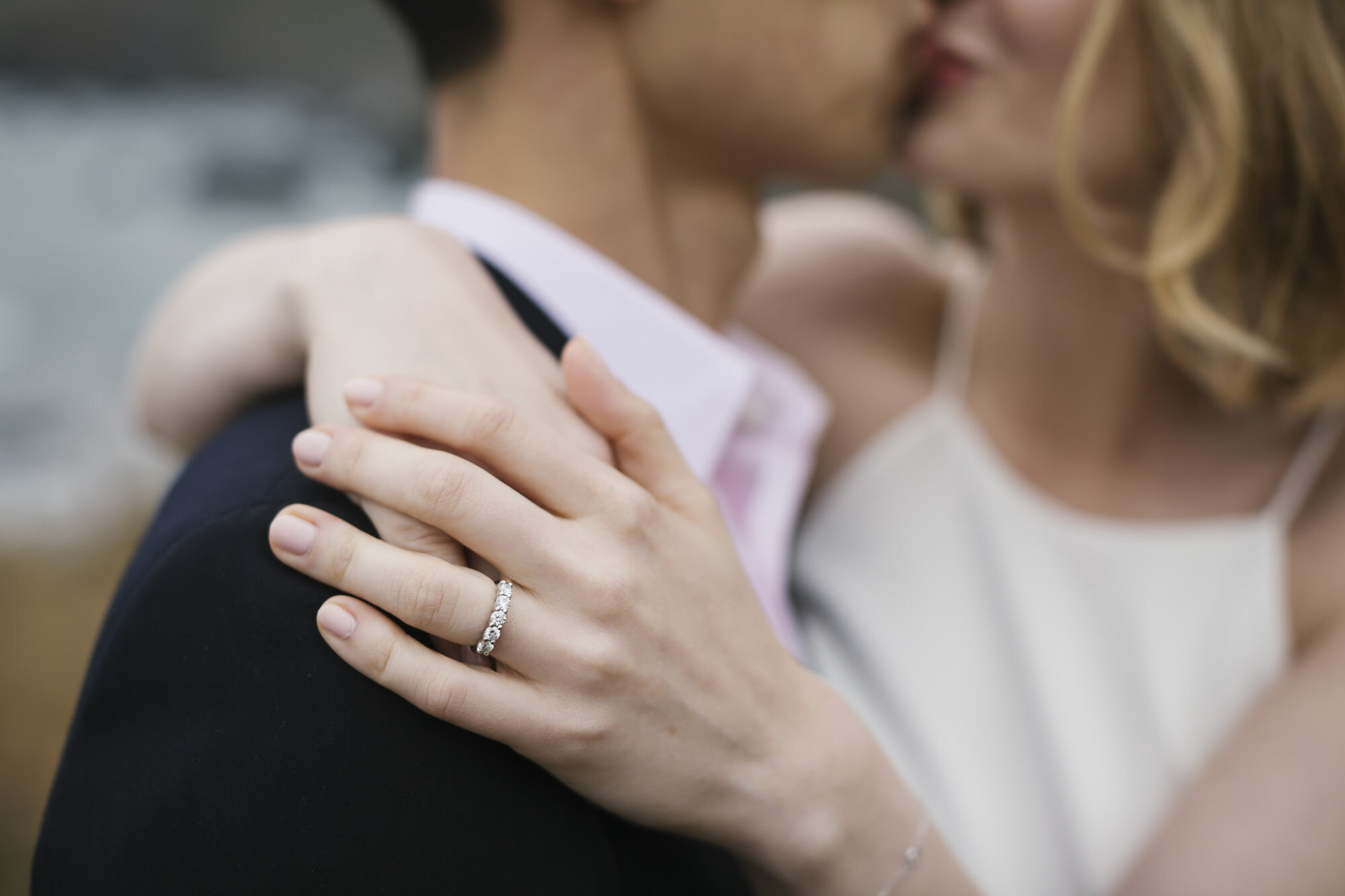 A close up of a woman's wedding ring on her hand while couple kiss in the background