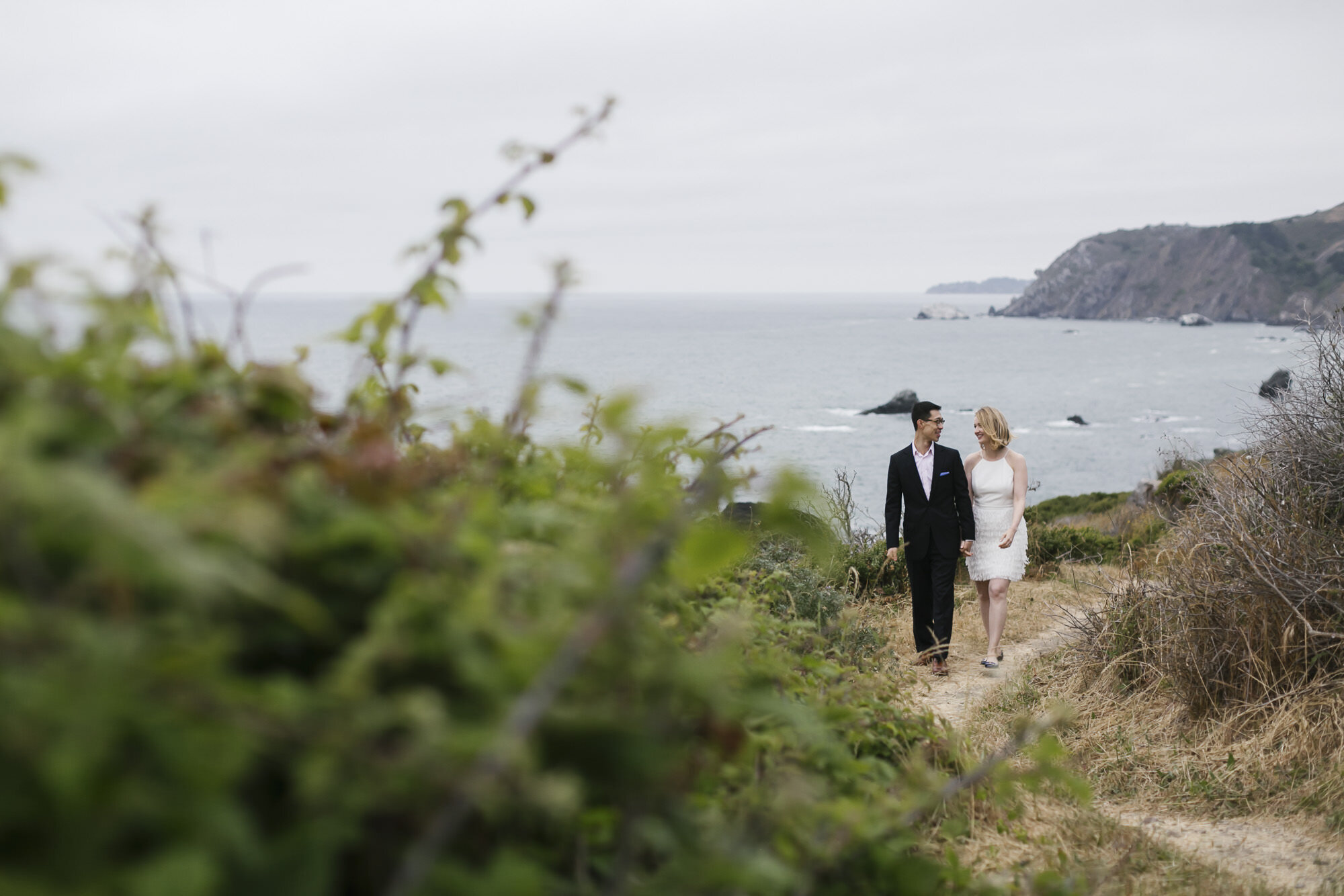 Wedding couple walk holding hands along hiking trail by the ocean