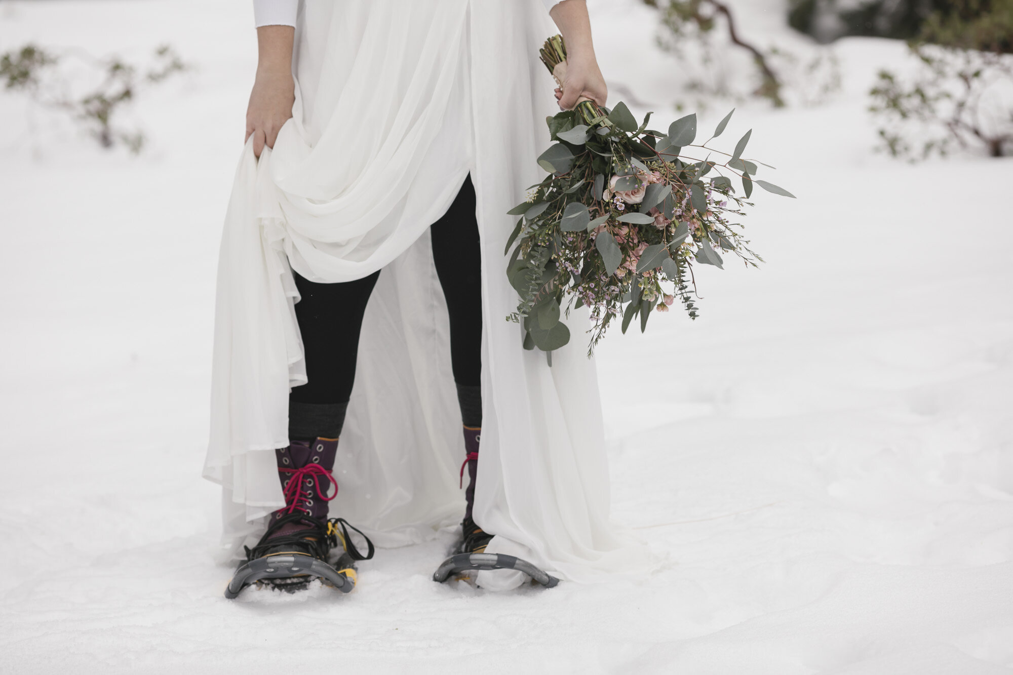 Winter bride shows off her hiking boots and snowshoes while holding her wedding bouquet