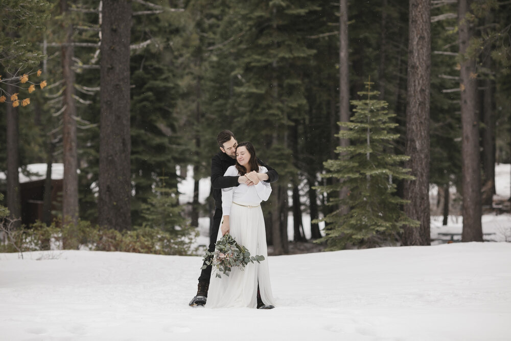 Groom hugs his bride from behind on their elopement day in the snow in Lake Tahoe