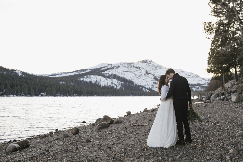 Bride and groom hold each other close alongside Lake Donner with the snowy summit behind them