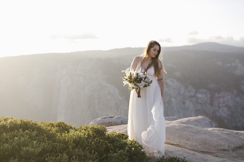 Bride holding her white wedding bouquet stands on Taft Point in Yosemite