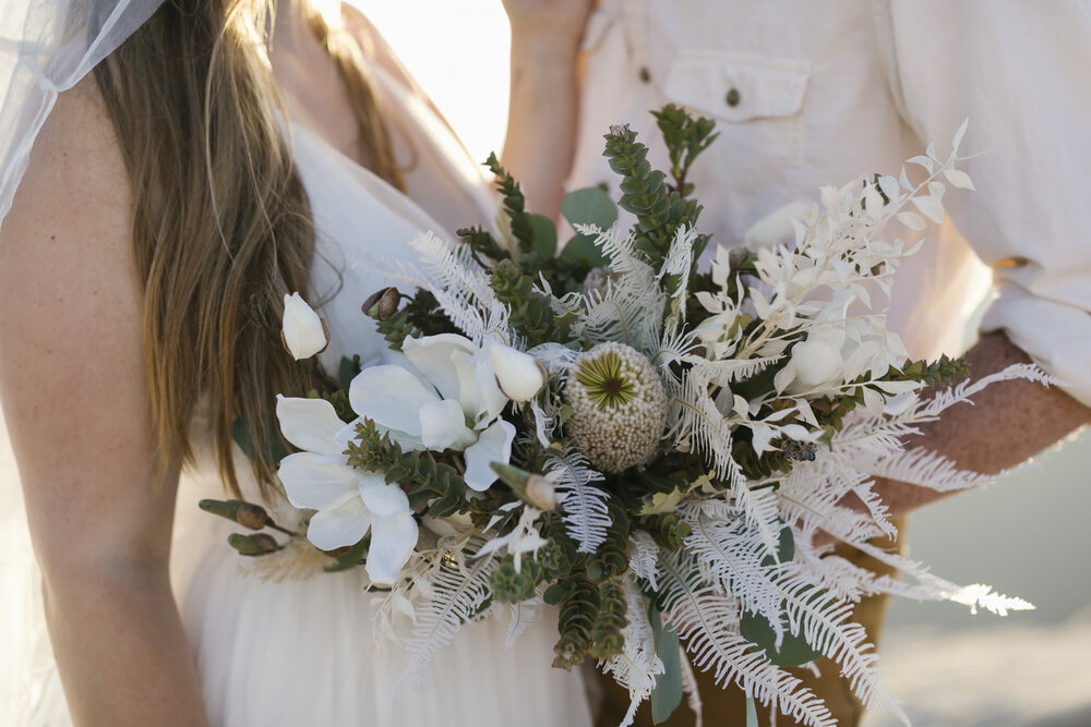 Groom holds bride's wedding bouquet made with eucalyptus and dried white flowers and ferns made by Soulflower