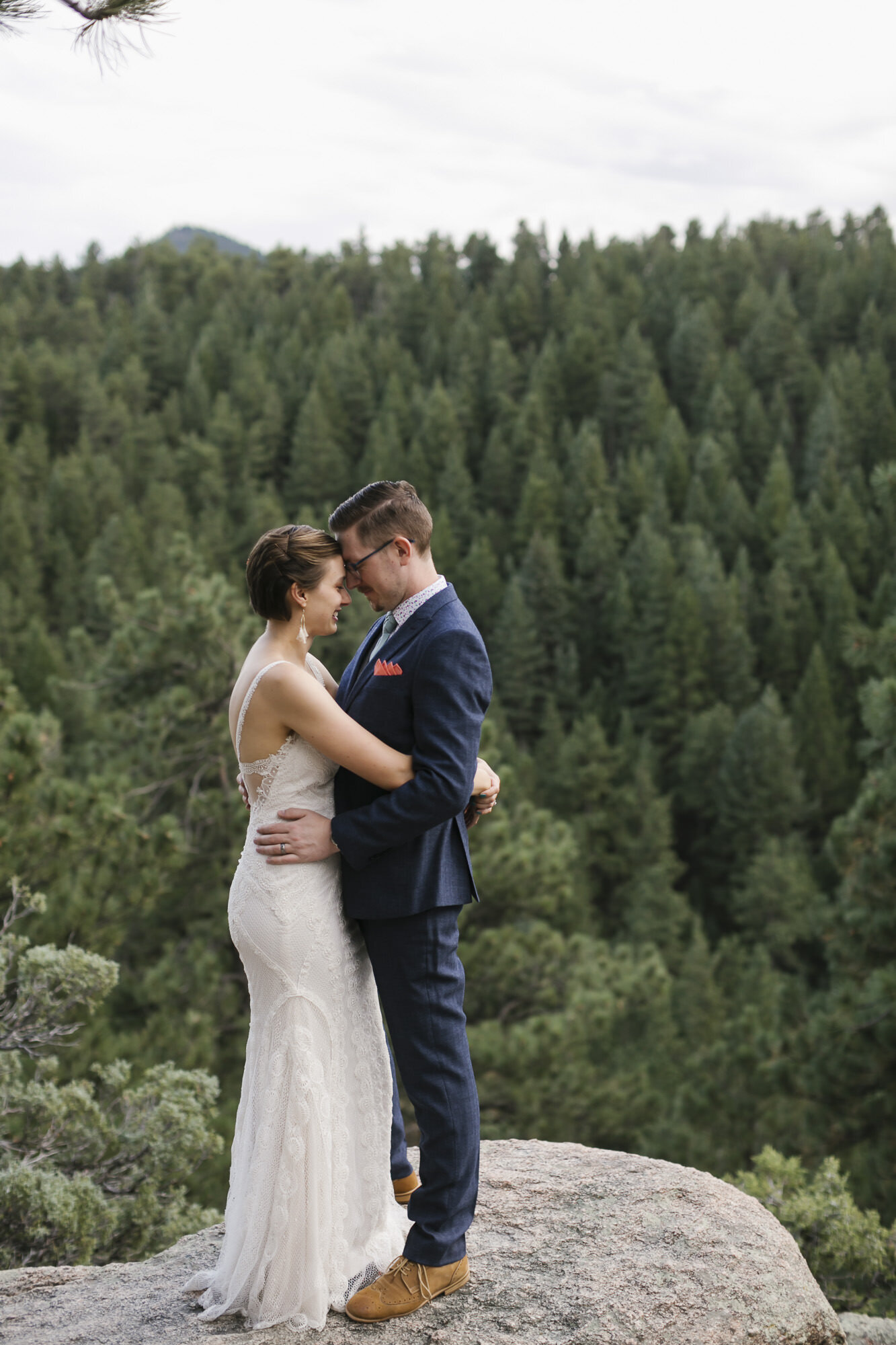 Wedding couple stand on rock outcropping with forest of trees behind them in Colorado