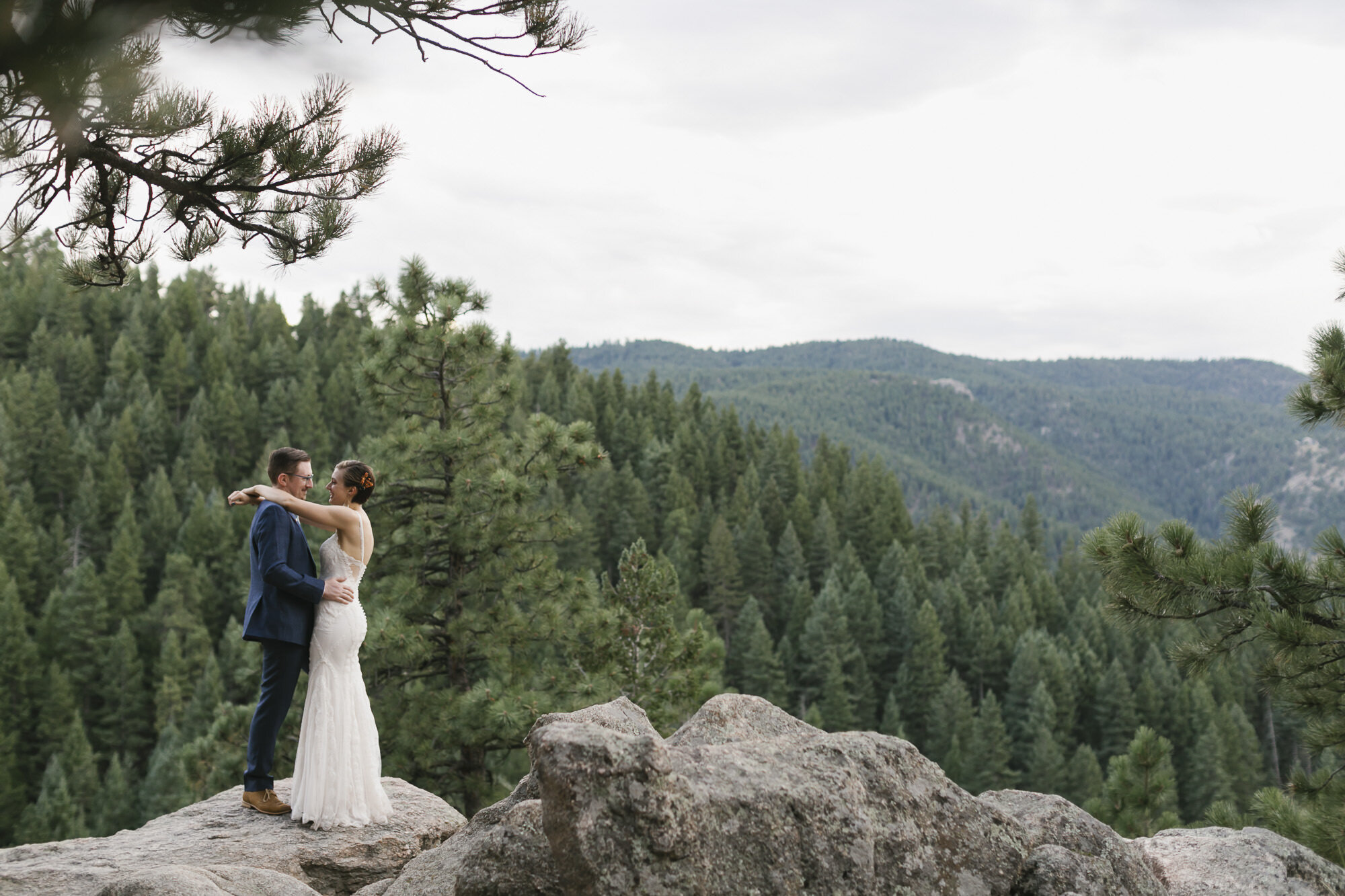 Bride and groom stand on rocky overlook with a pine forest in the background