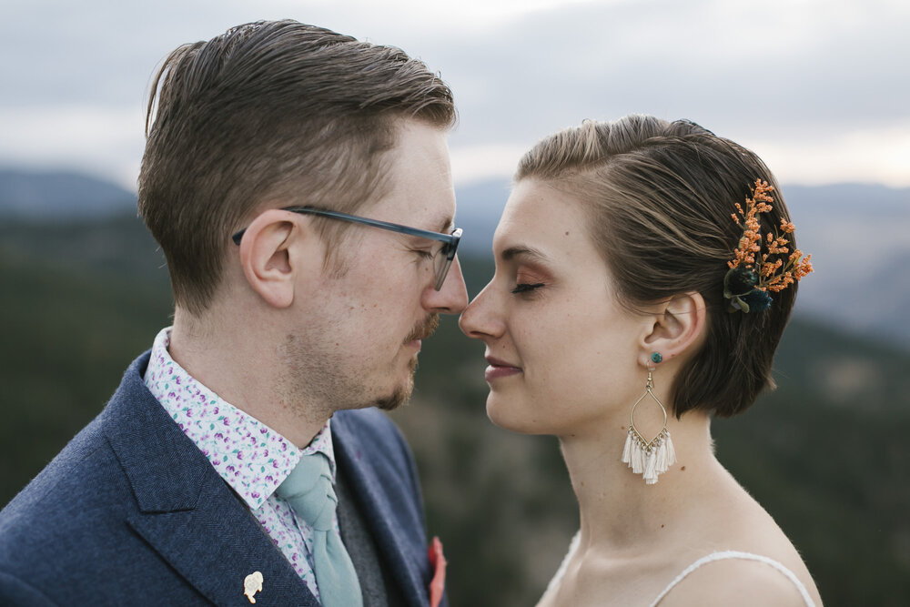 Bride with short hair, white fringe earrings, and orange combs with her groom in a blue suit with brightly patterned shirt snuggle close