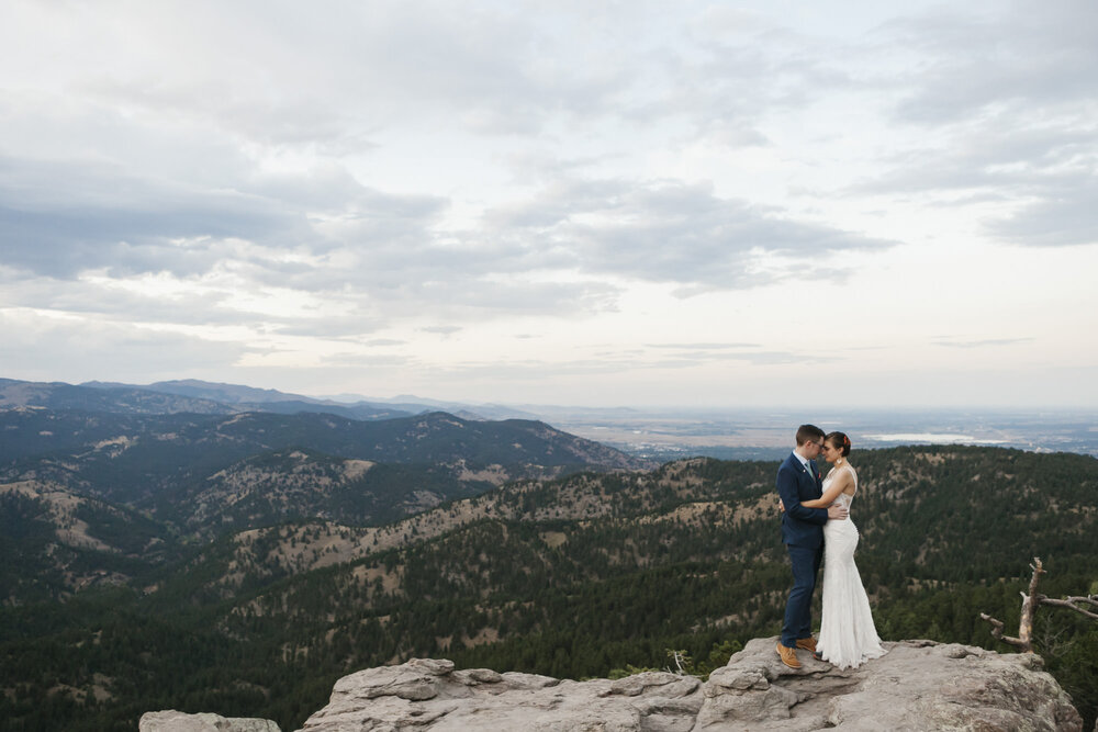 Wedding couple stand on rocky overlook with mountains all around them above Boulder Colorado