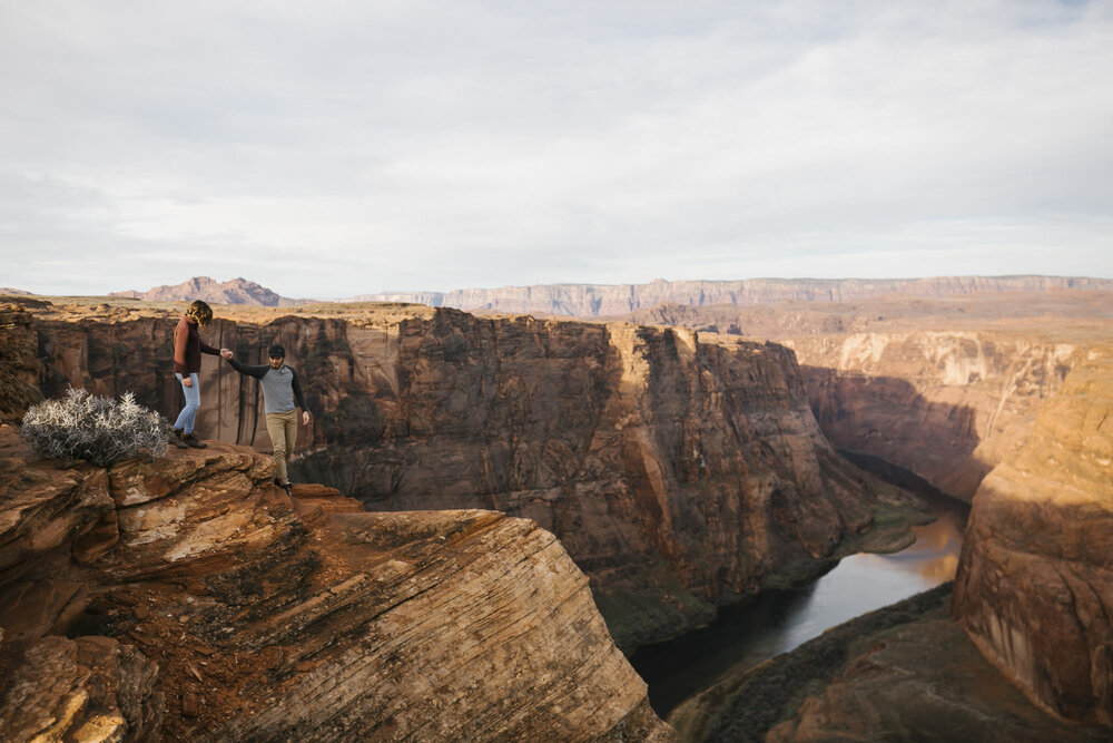 Engaged couple help each other walk along the rim at Horseshoe Bend in Arizona at sunrise