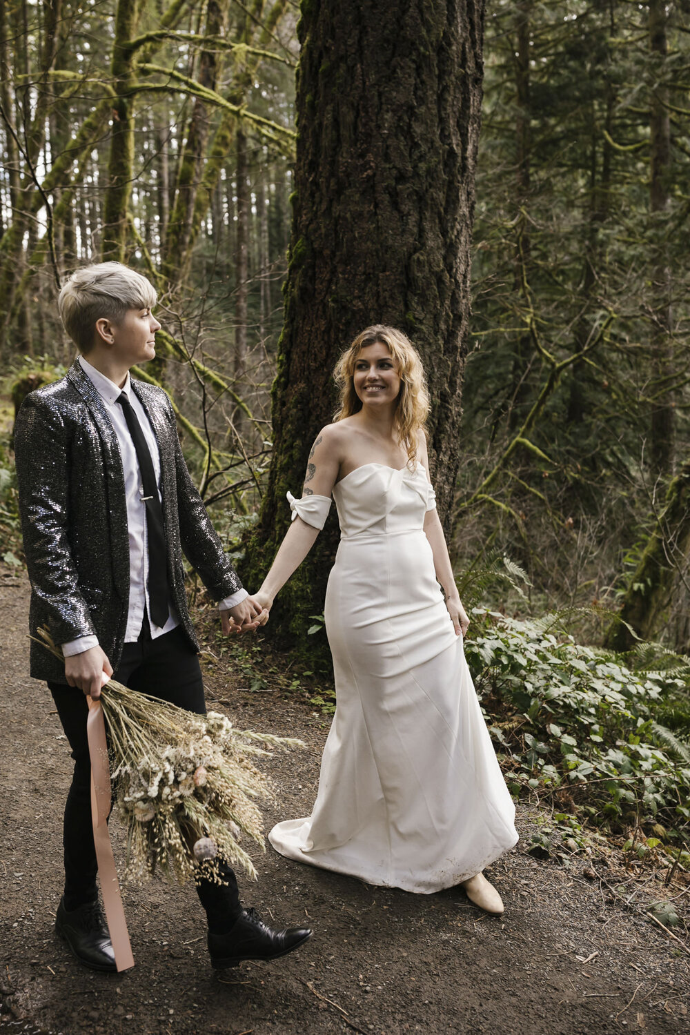 Same sex couple, one wearing a white wedding dress, the other wearing a sequined jacket walk in a Washington forest for their elopement in the PNW