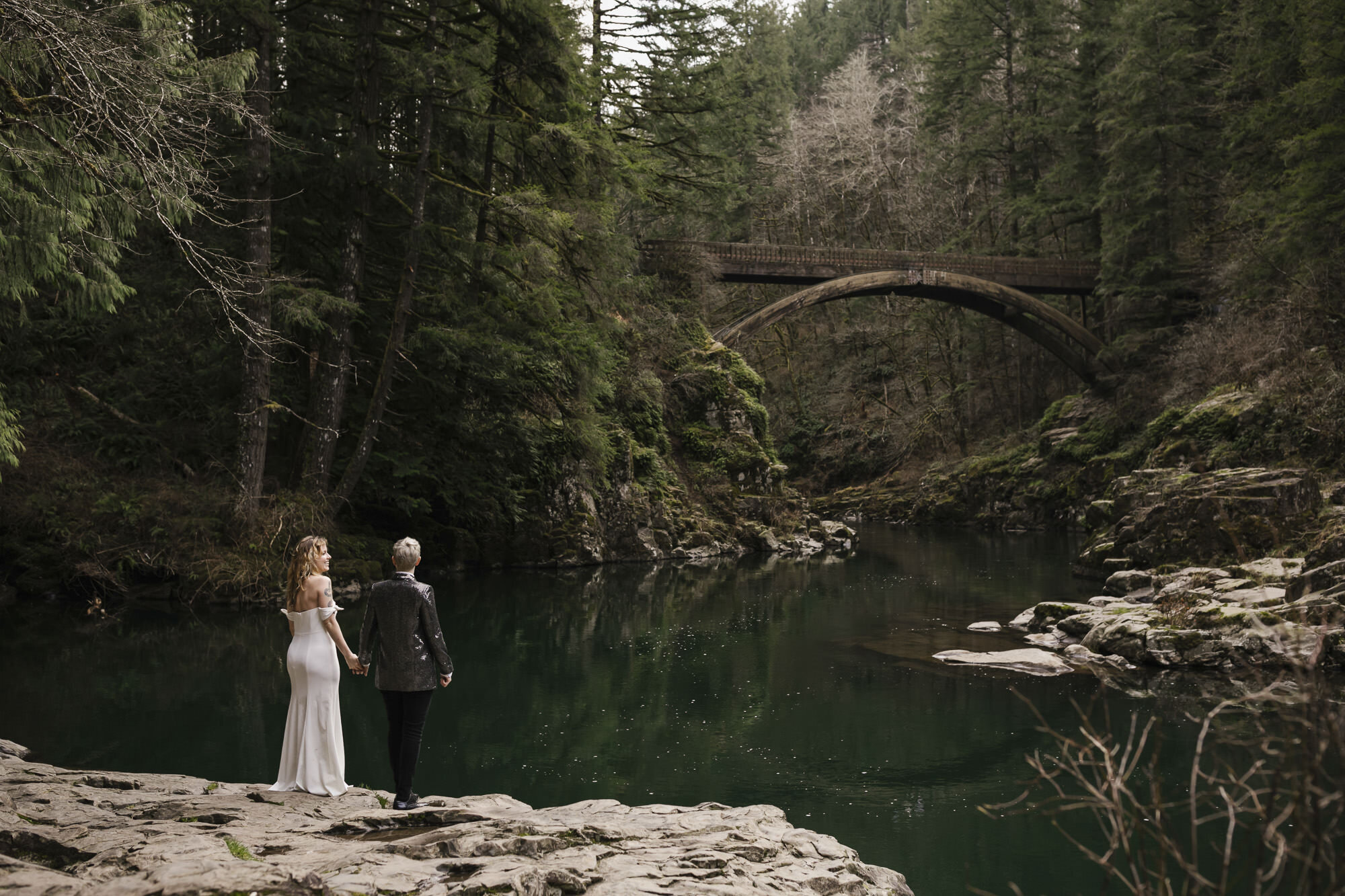 Same sex couple stand together in front of the bridge at Moulton Falls in Washington