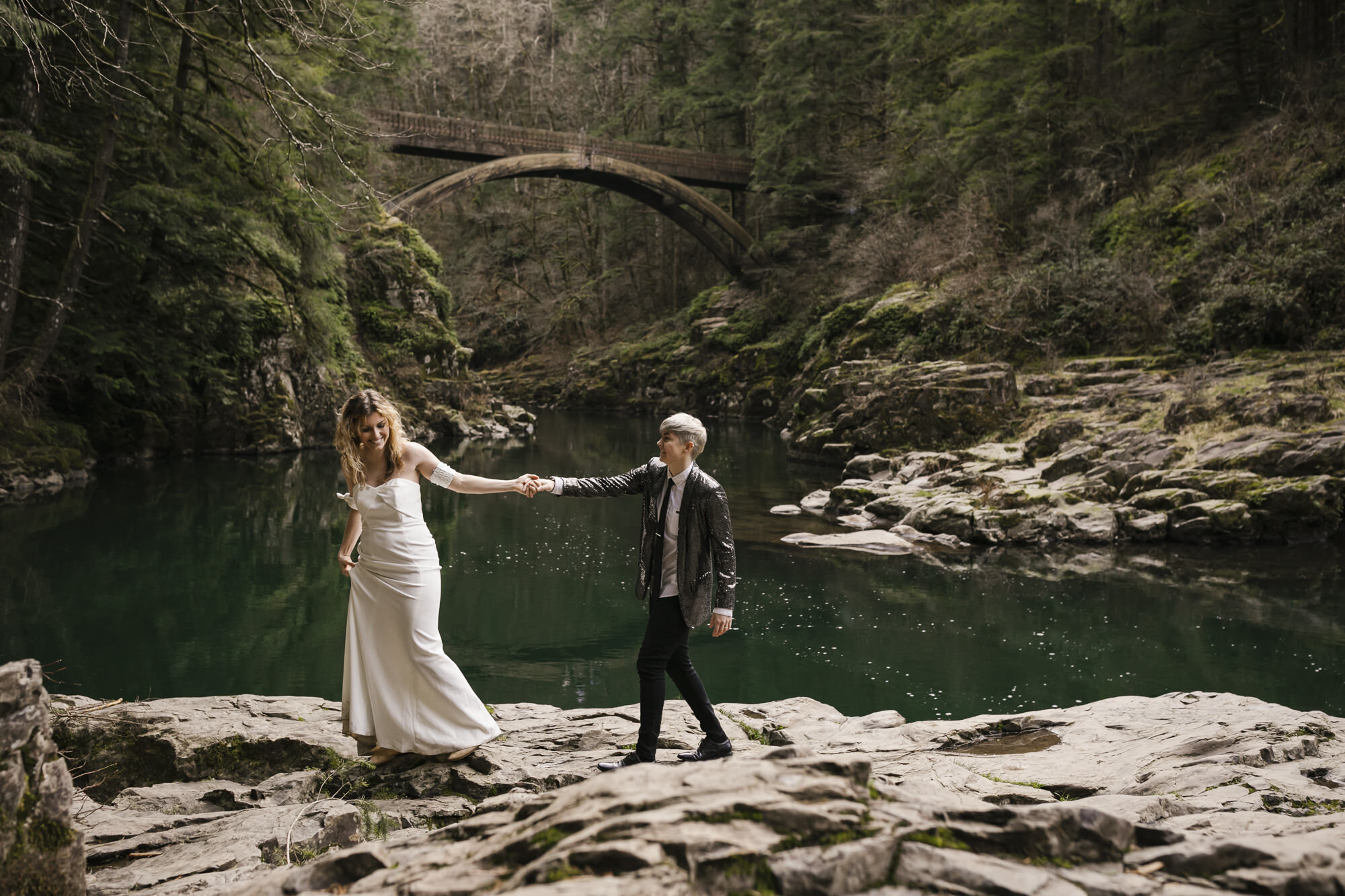 Bride playfully leads her partner walking across the rocks in front of the bridge at Moulton Falls in Washington