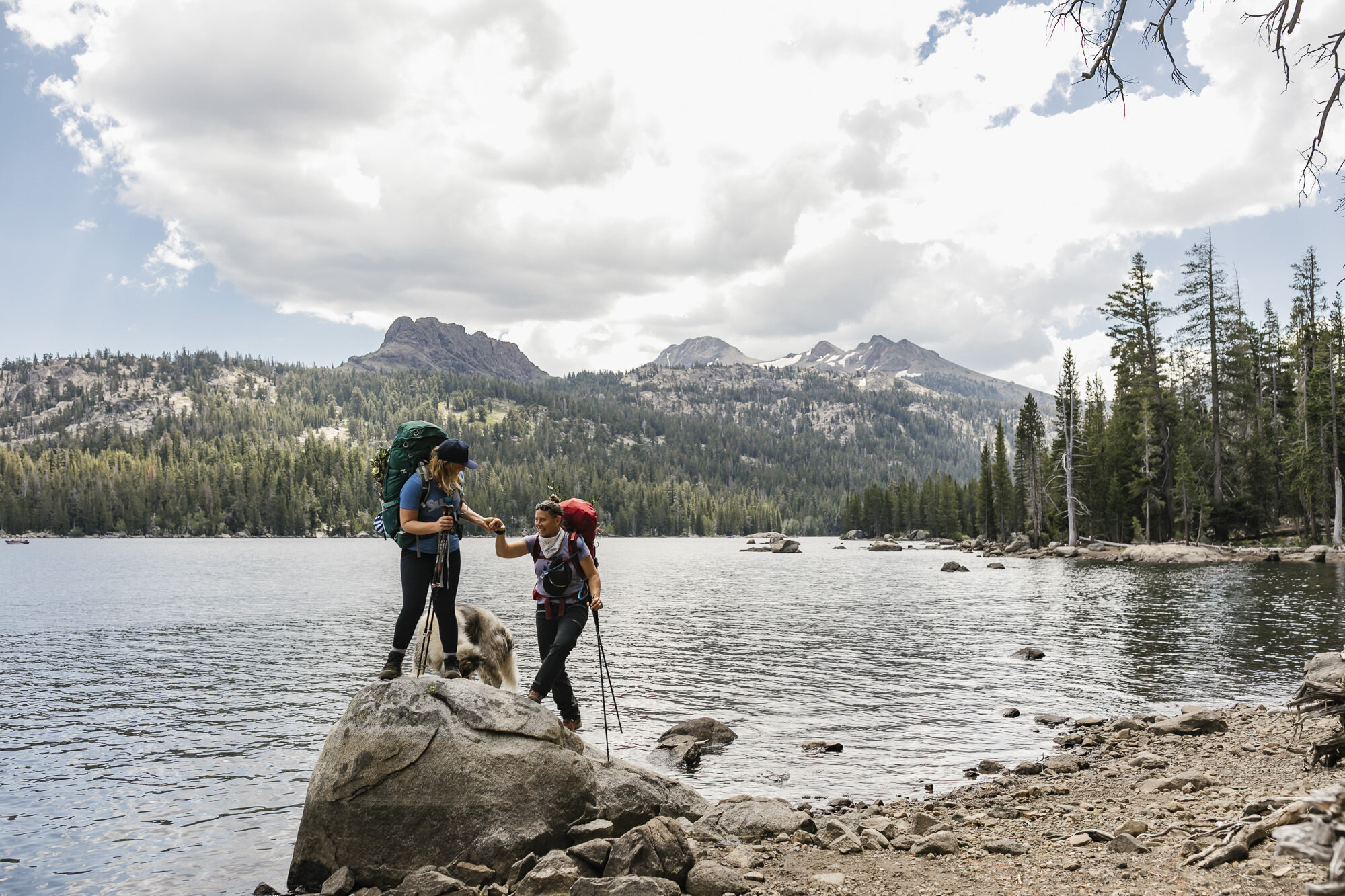 Elopement couple help each other climb rock with their dog in front of an alpine lake in the Sierra Nevada mountains