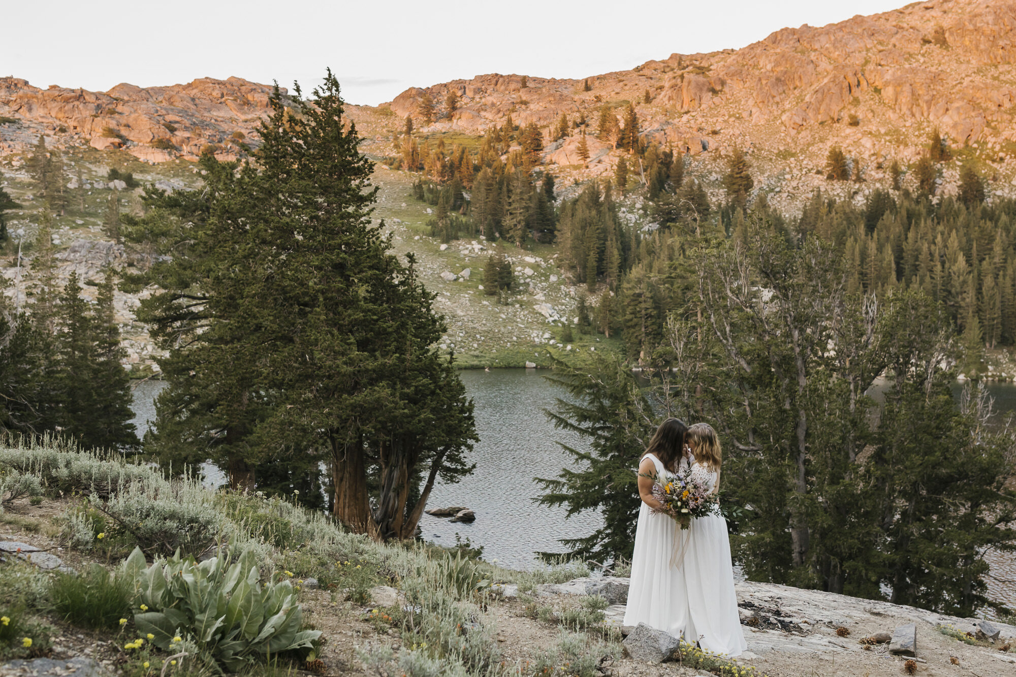 Same sex wedding couple on their wedding day stand in front of an alpine lake with alpenglow on the mountains behind them