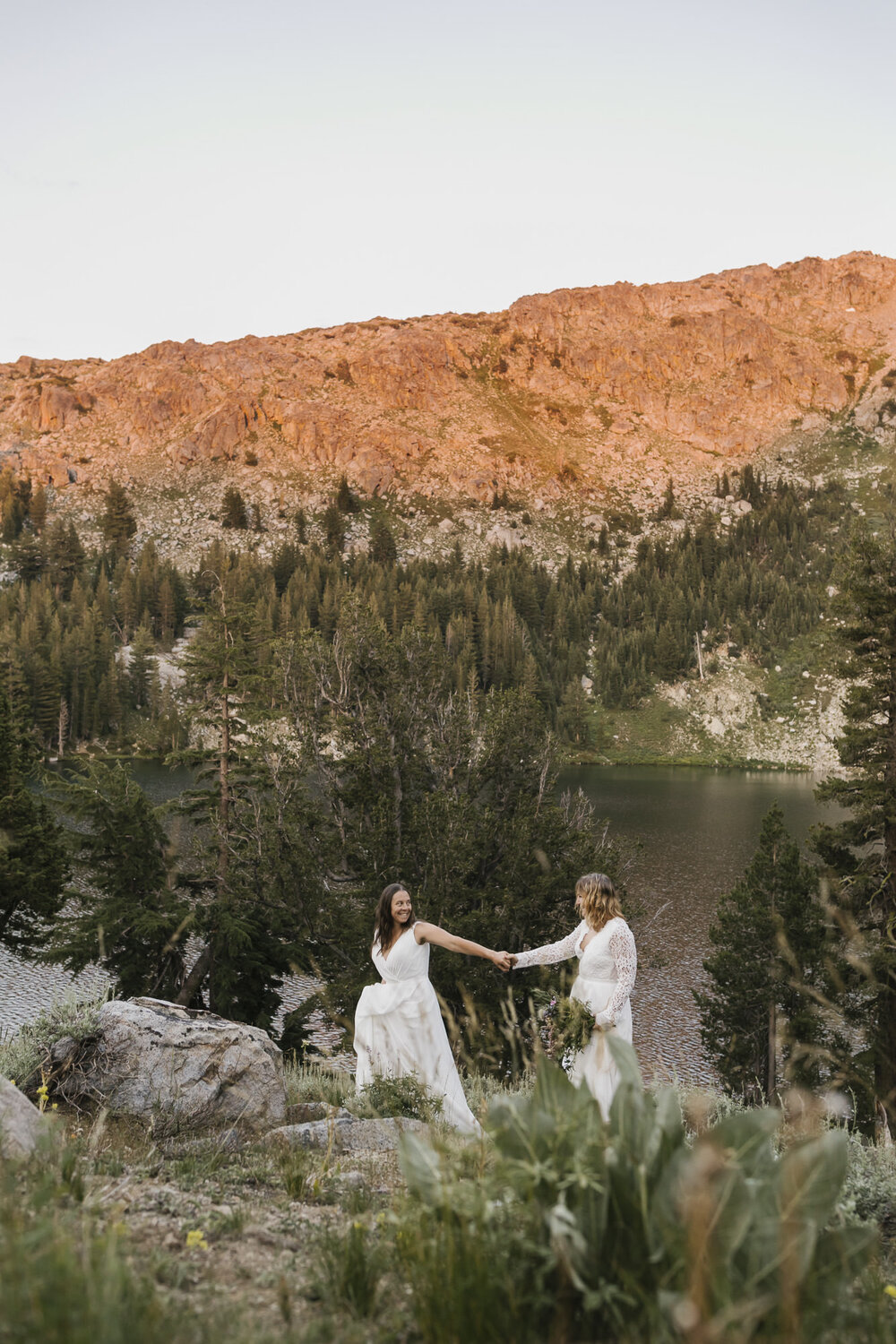 Two brides walk holding hands during their backpacking elopement with alpenglow on the mountains behind them
