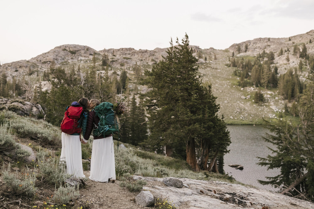 Wedding couple in their dresses, puffy jackets, and backpacks, kiss in front of an alpine lake during their mountain elopement