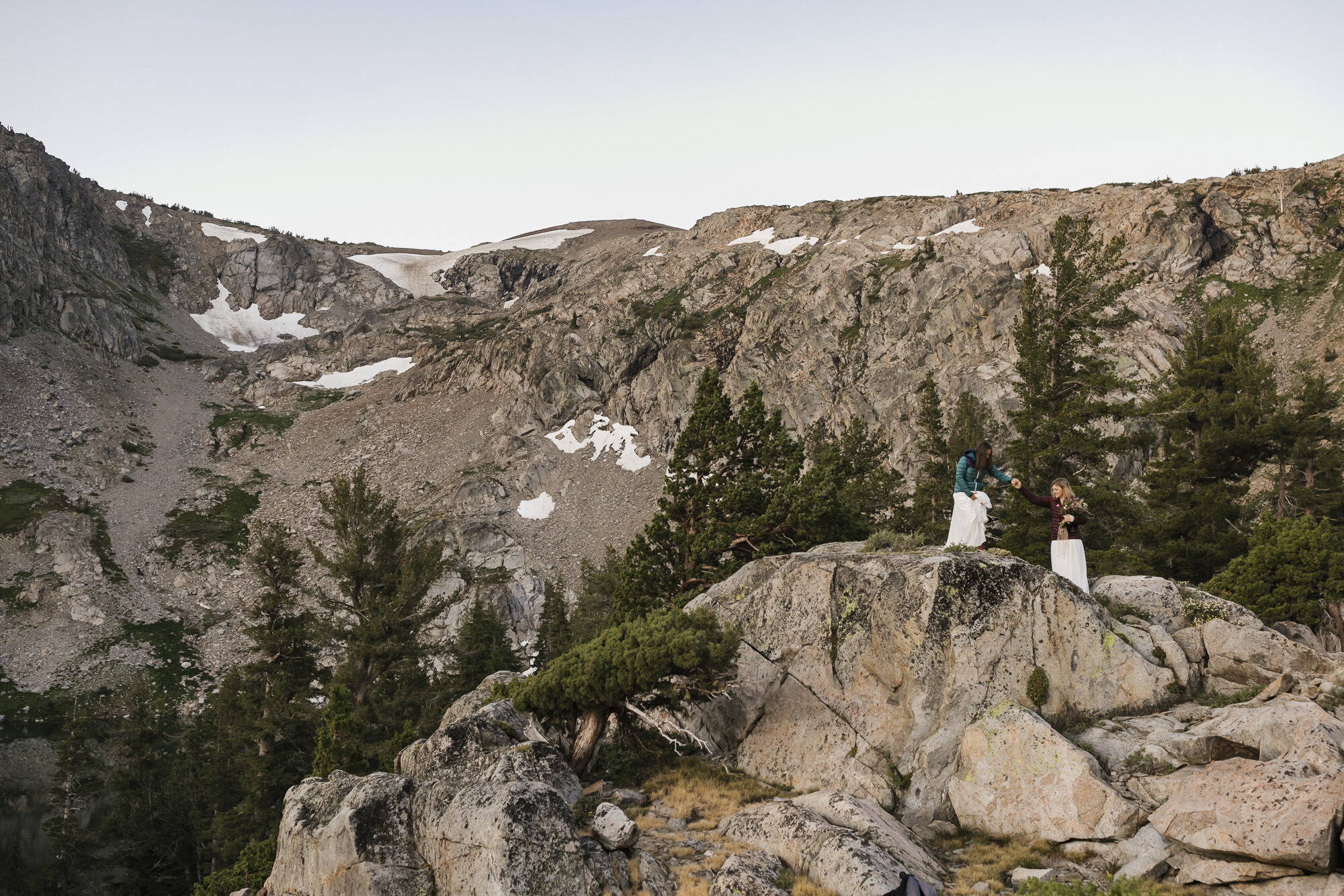 Wedding couple help each other over rocks in the Sierra Nevada mountains at sunrise during their backpacking elopement