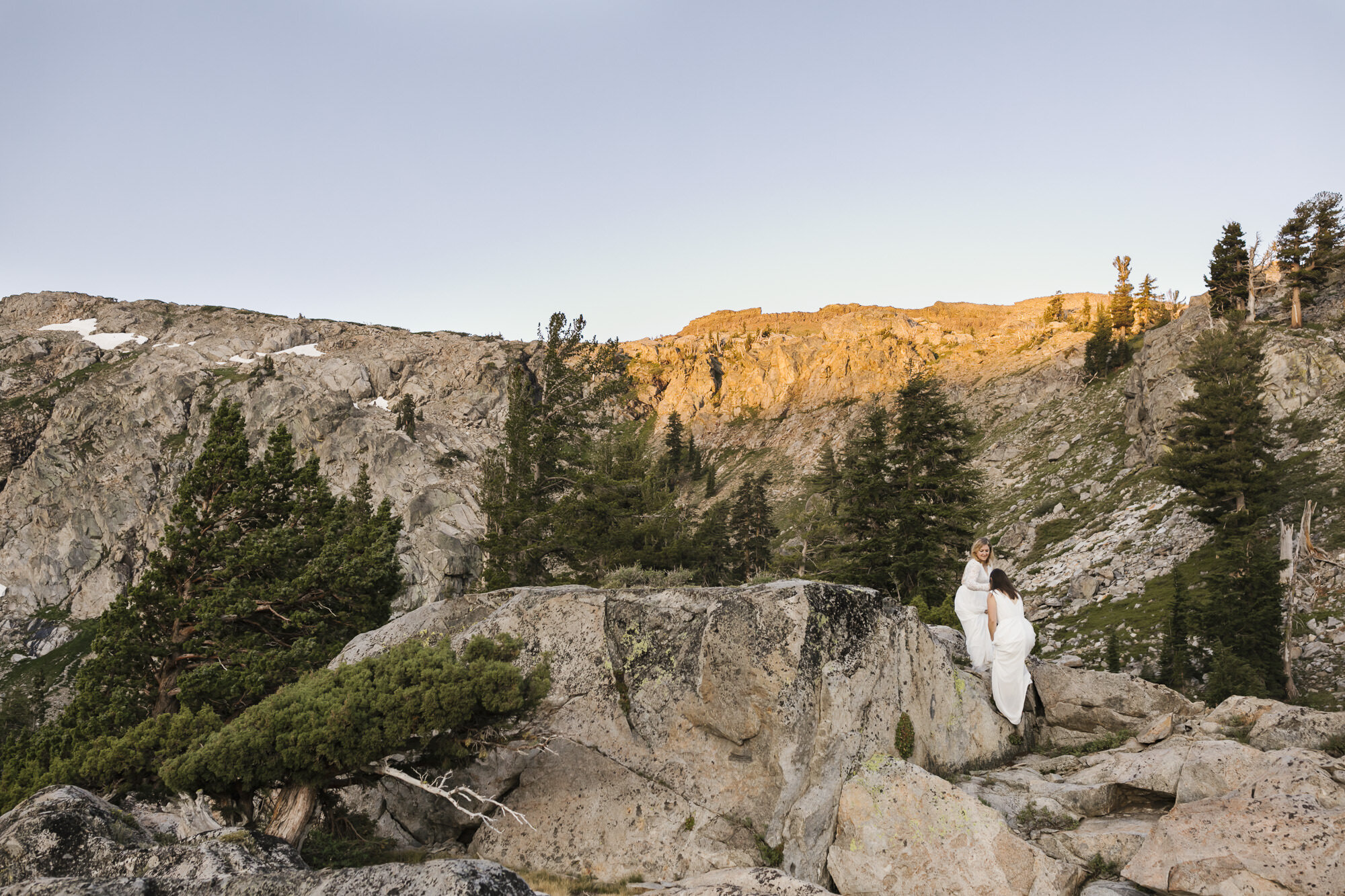 Wedding couple help each other climb rocks at sunrise with the alpenglow behind them in the California mountains