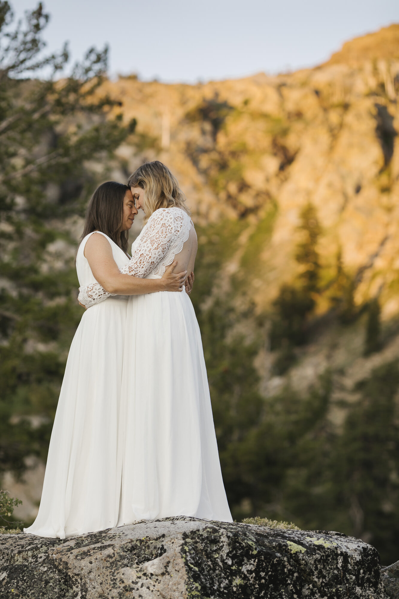 Same sex wedding couple hold each other close with sunrise alpenglow on the mountains behind them
