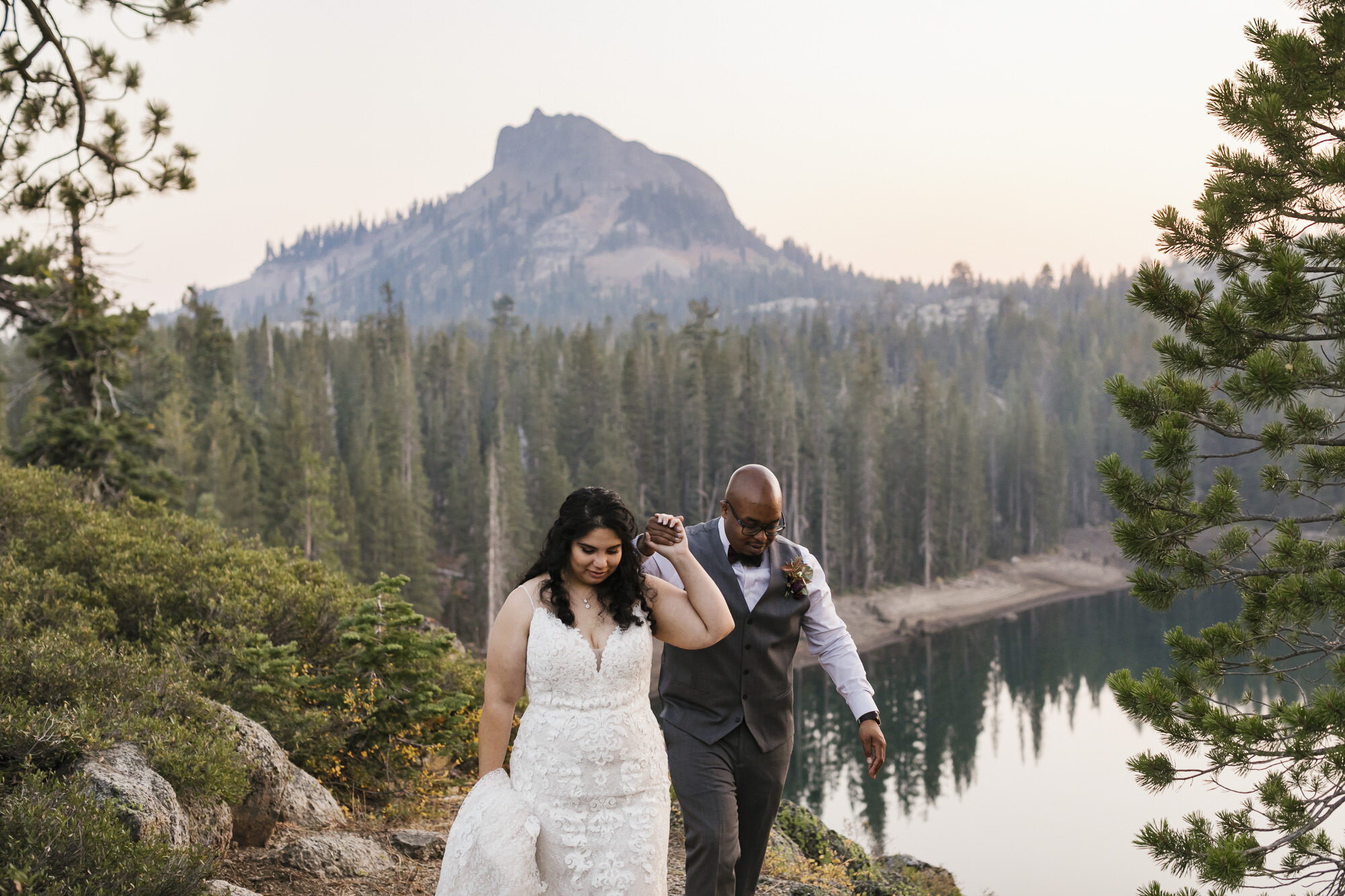 Wedding couple walk holding hands with mountain peak behind them in Tahoe forest