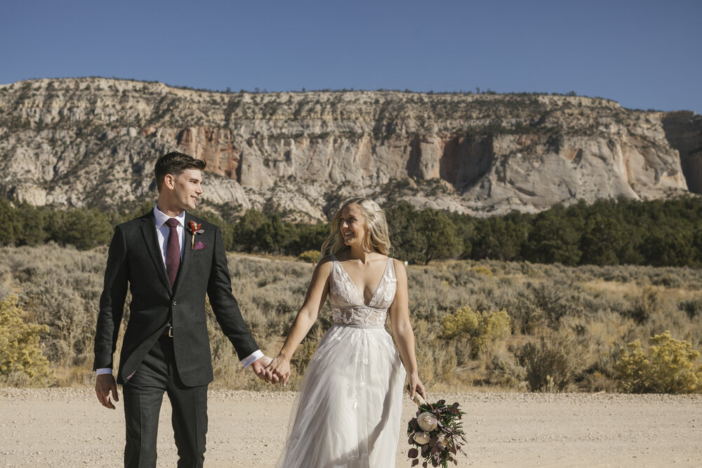 Wedding couple walk together holding hands in front of the red cliffs at their Utah Airbnb outside of Zion