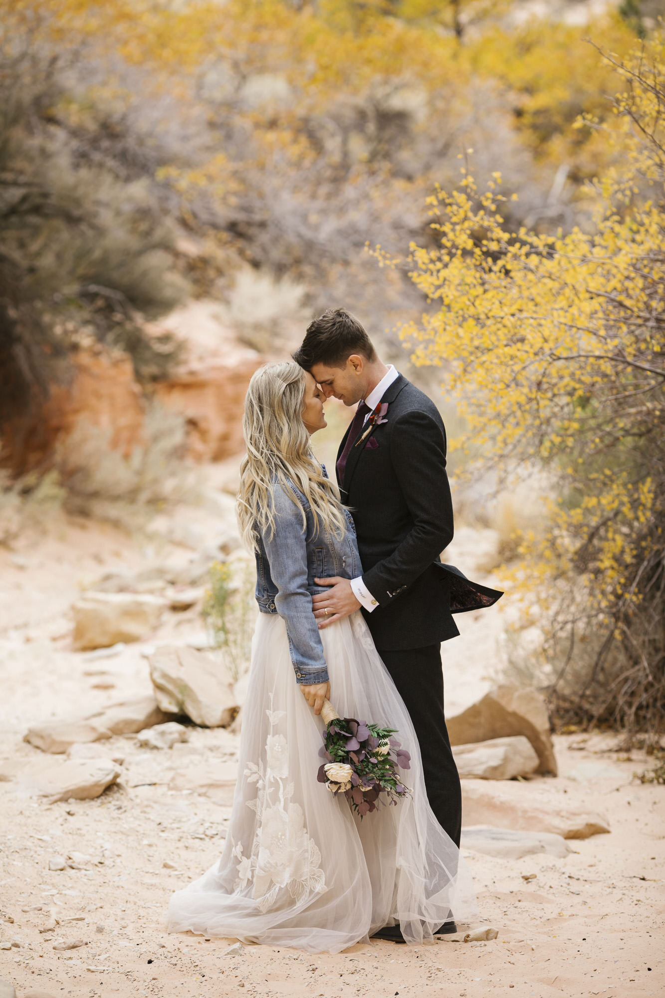 Bride in denim jacket stands with her groom during their day-after wedding portraits in the Utah desert