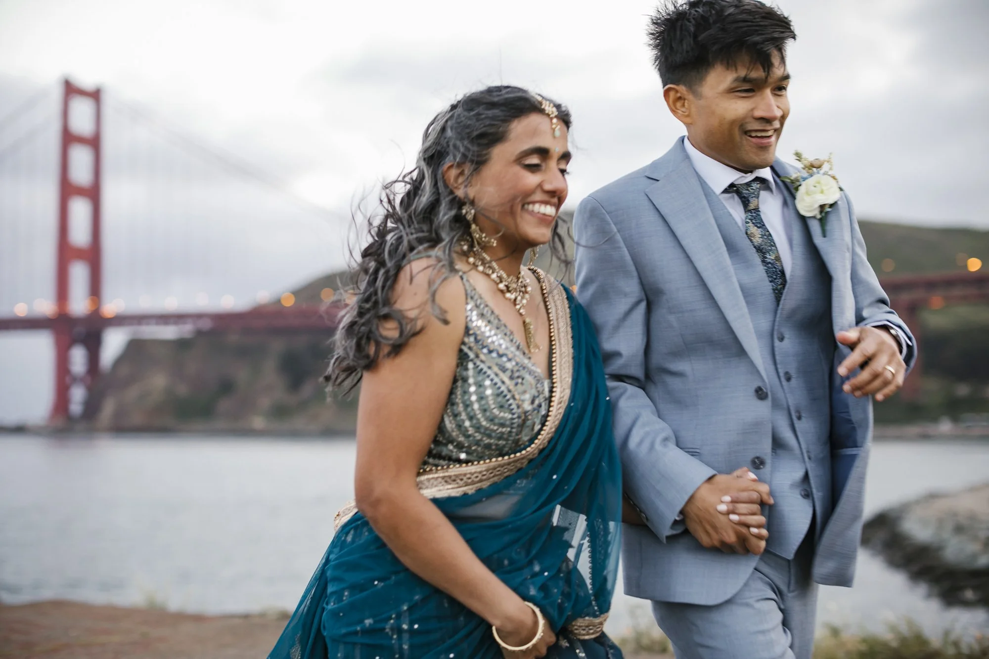 A bride wearing a blue saree and her husband in a grey three piece suit hold hands in front of the Golden Gate bridge in Sausalito