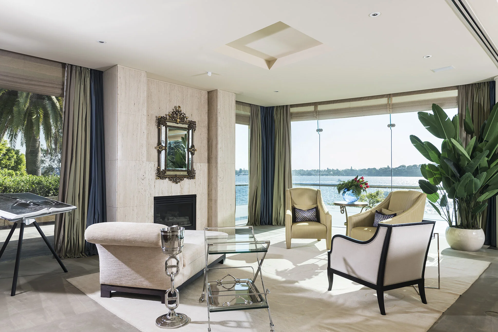 Luxury sitting room at Watkins Residence featuring bespoke furnishings, natural stone fireplace, floor-to-ceiling glazing and panoramic waterfront views, creating an elegant retreat connected to its riverside setting.