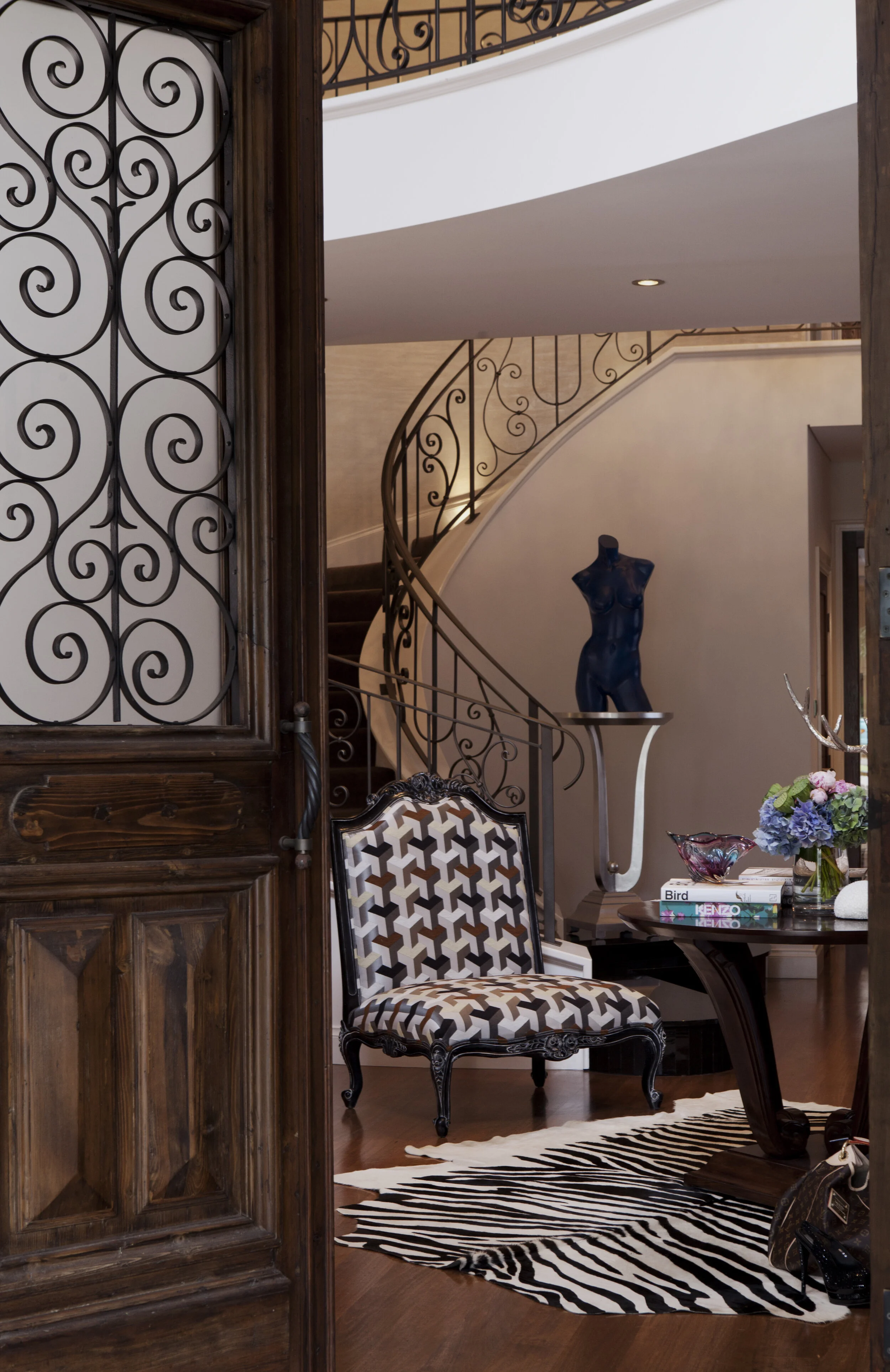 Luxury residential entry foyer featuring carved timber doors, geometric upholstered accent chair, zebra hide rug, sculptural staircase and curated designer styling.