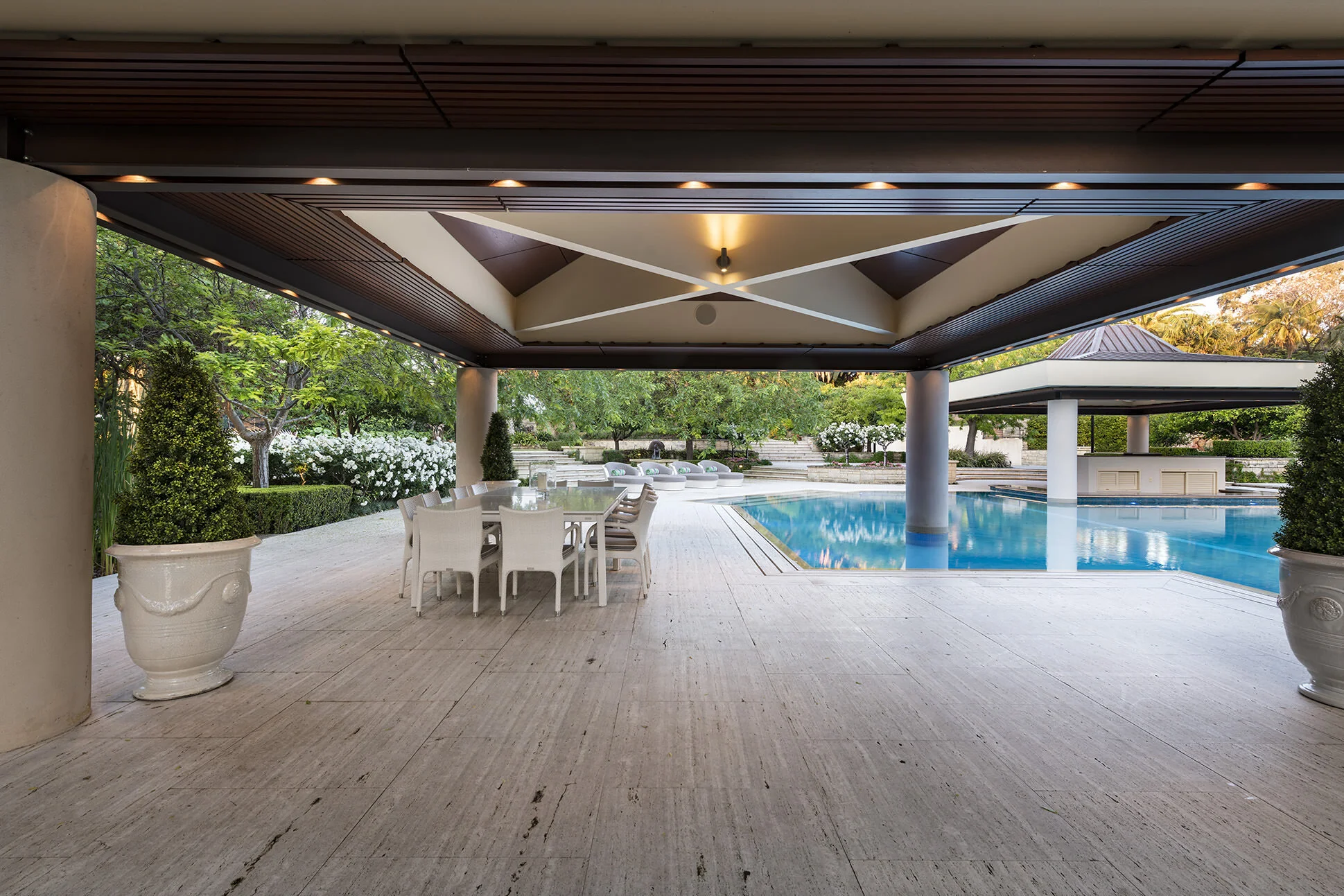Covered outdoor entertaining pavilion at Watkins Residence featuring a resort-style dining area, swimming pool and landscaped gardens, designed for luxury indoor-outdoor living.