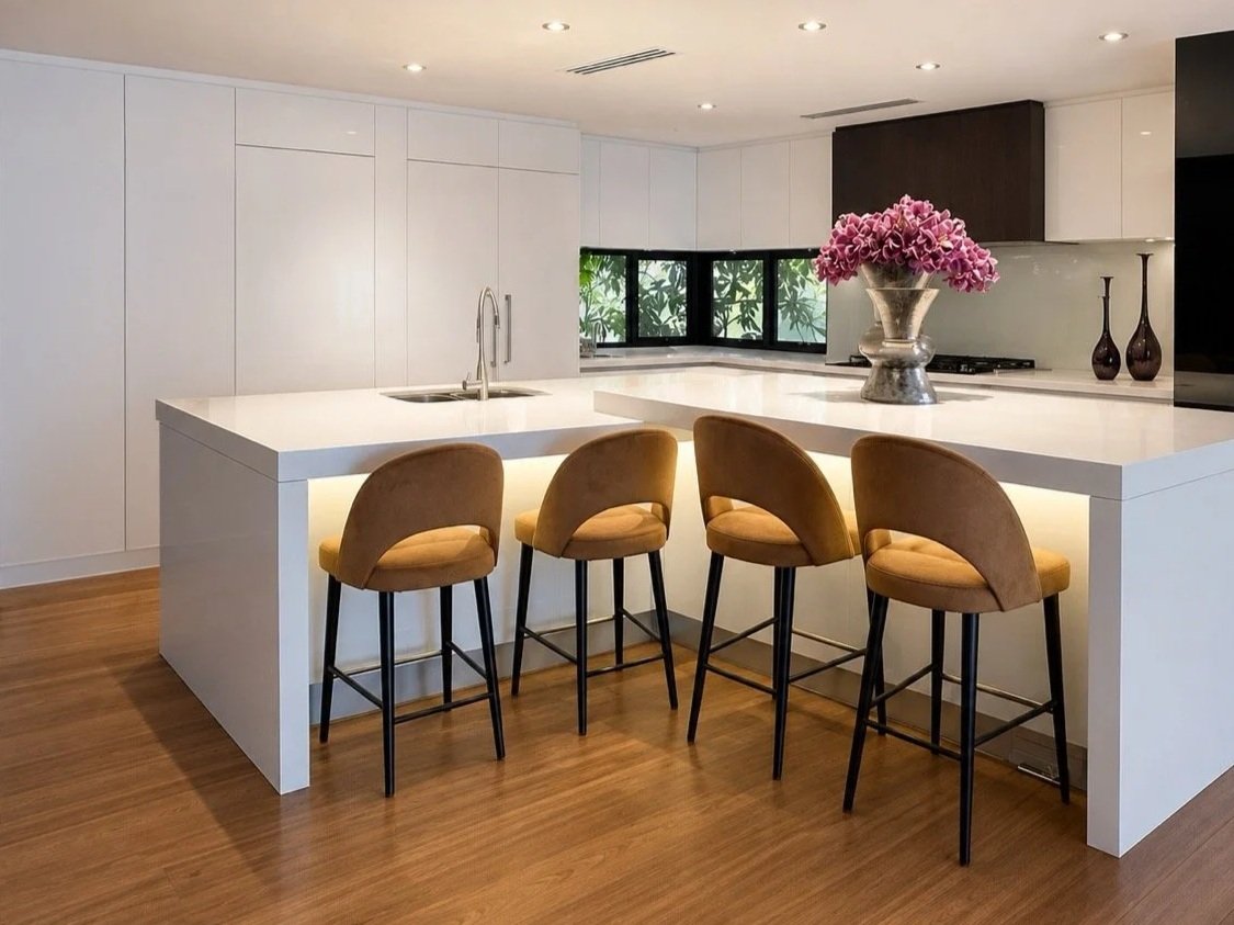 Kenmore Residence kitchen with white cabinetry, large island bench, tan bar stools, timber flooring and pink floral arrangement.