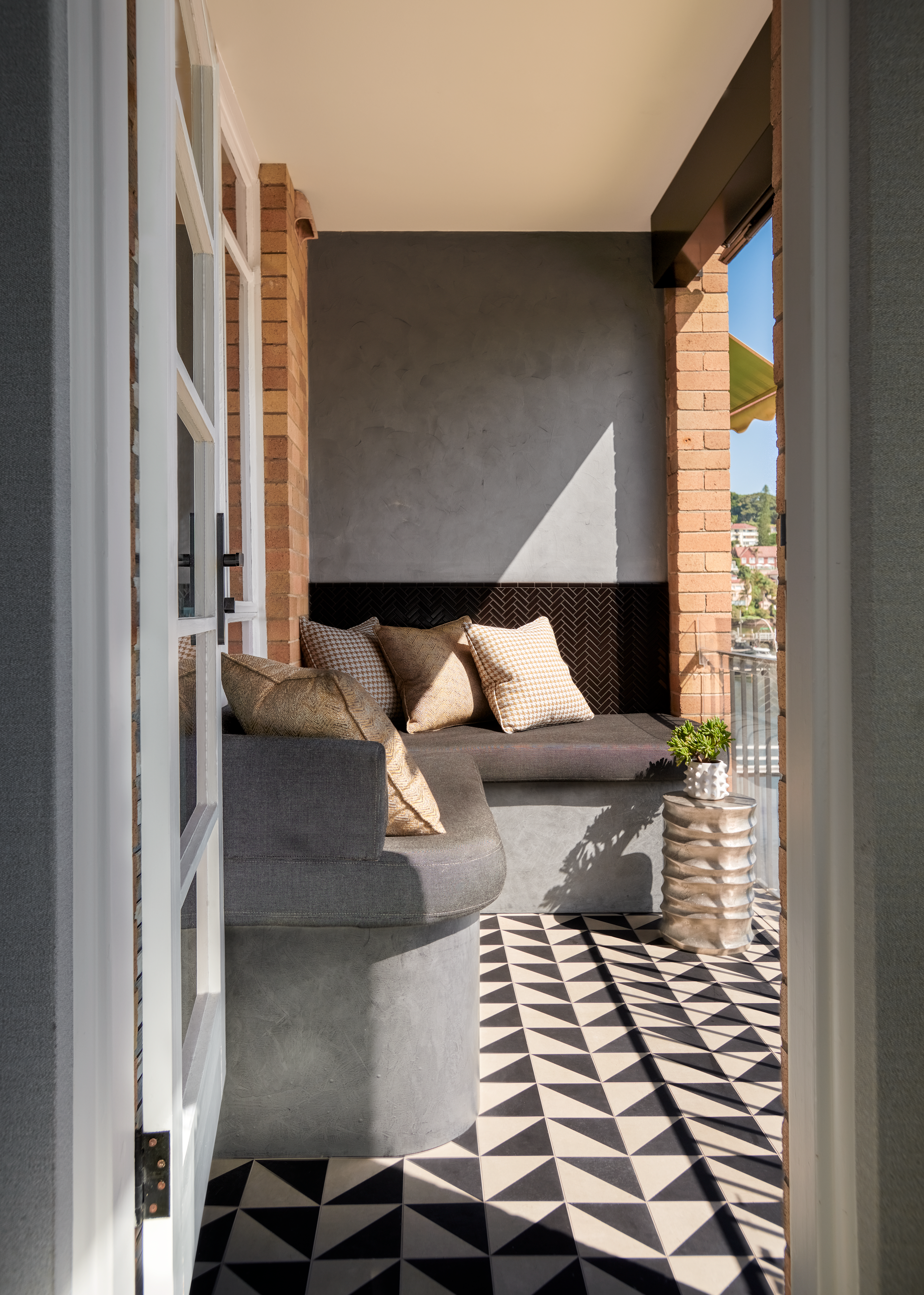 Covered balcony retreat with built-in curved banquette seating, textured finishes and geometric floor tiles. Warm neutral tones, layered cushions and filtered natural light create an intimate outdoor living space with architectural character.