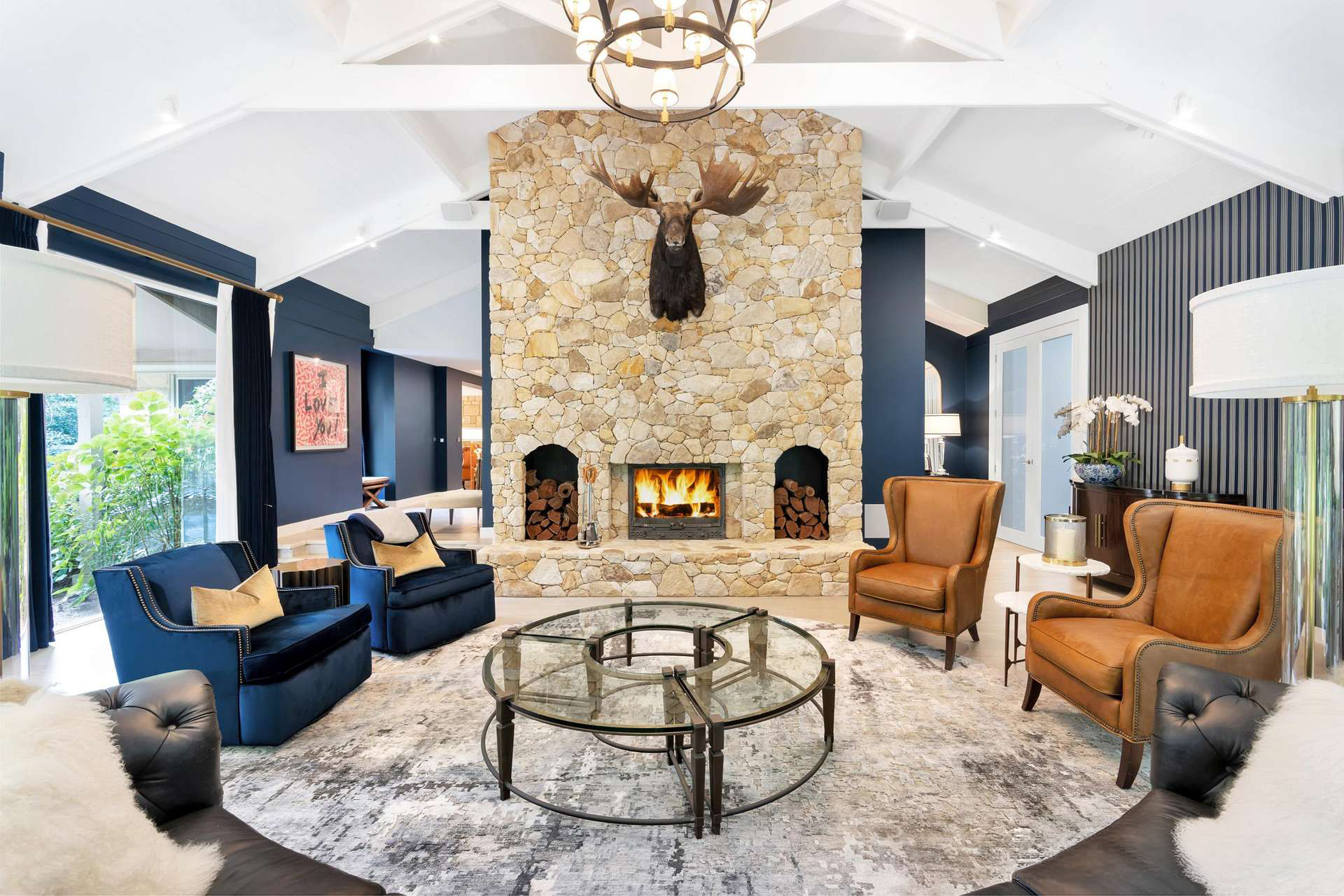 Luxury living room designed by Luis La Pegna featuring a grand stone fireplace with mounted moose, vaulted white ceilings, navy wall panelling, leather wingback chairs, custom seating, and contemporary furnishings in an elegant residential interior.
