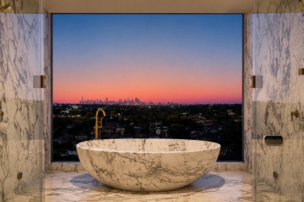 Burwood Penthouse master ensuite featuring a freestanding marble bathtub, marble-lined walls, brass tapware, glass shower screens and expansive sunset city views.