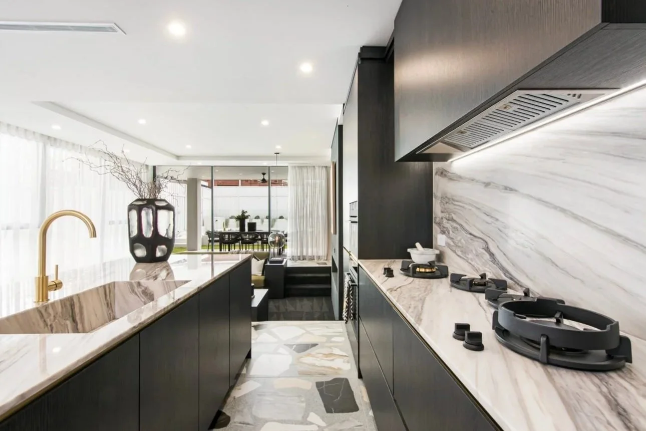 Luxury kitchen at Wolli Residence featuring dark custom cabinetry, dramatic marble benchtops and splashback, integrated appliances, brass tapware and terrazzo flooring, creating a sophisticated contemporary entertaining space.