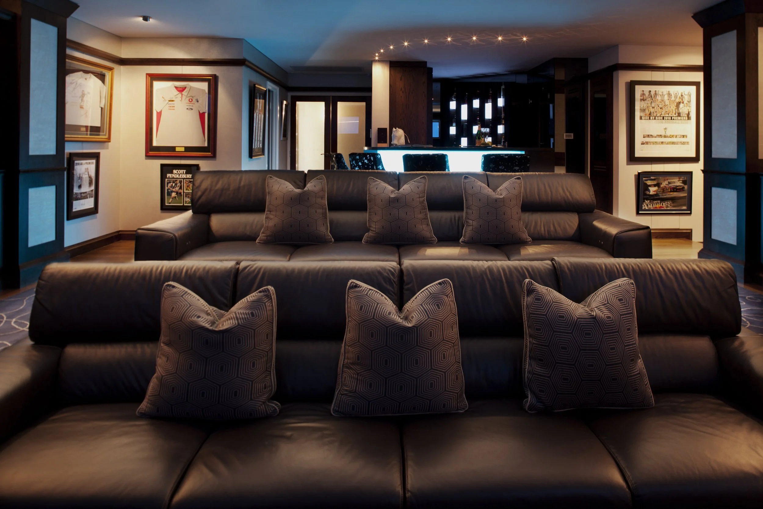 Kenmore Residence home theatre lounge featuring tiered leather seating, layered cushions, framed artwork, ambient blue lighting and a bar area in the background.
