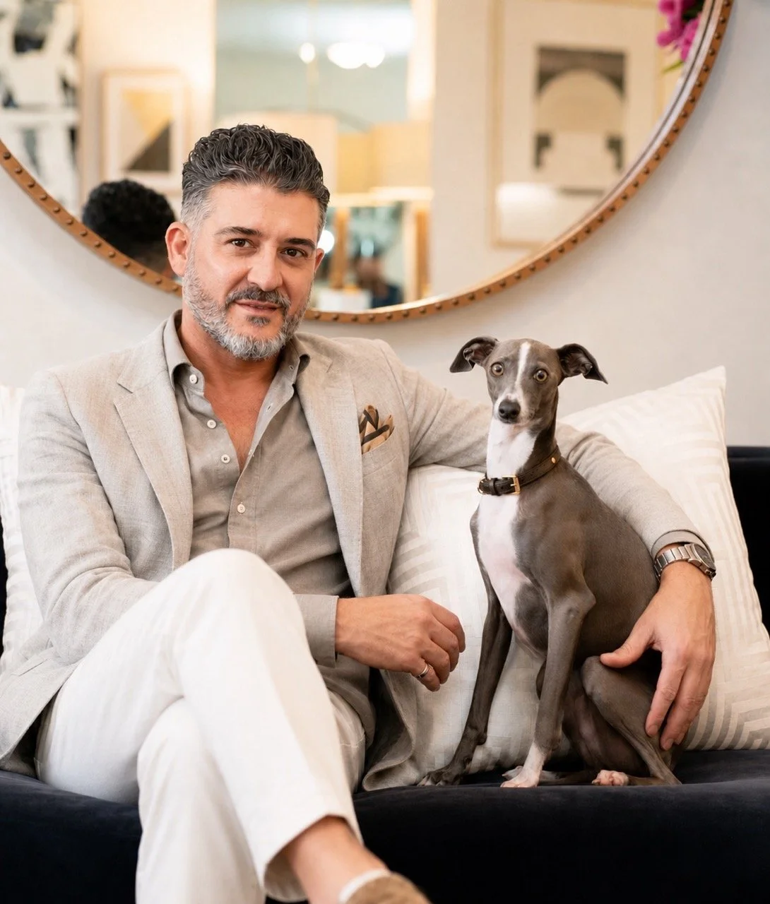 Portrait of interior designer Luis La Pegna seated on a navy sofa with his Italian Greyhound, featuring a tailored neutral outfit and warm contemporary interior backdrop.
