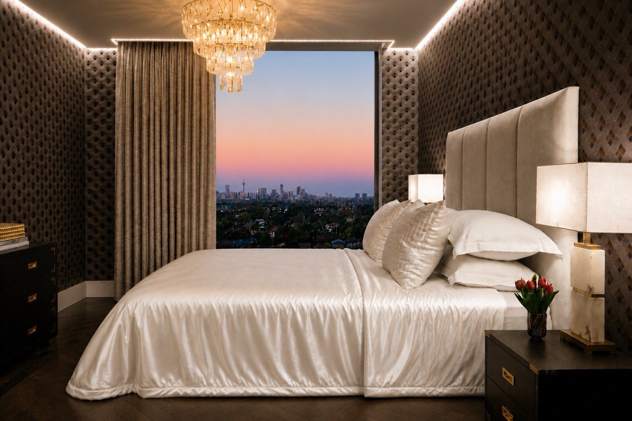 Burwood Penthouse master bedroom featuring a luxury bed with silk bedding, upholstered bedhead, patterned wall covering, crystal chandelier and sunset city views.