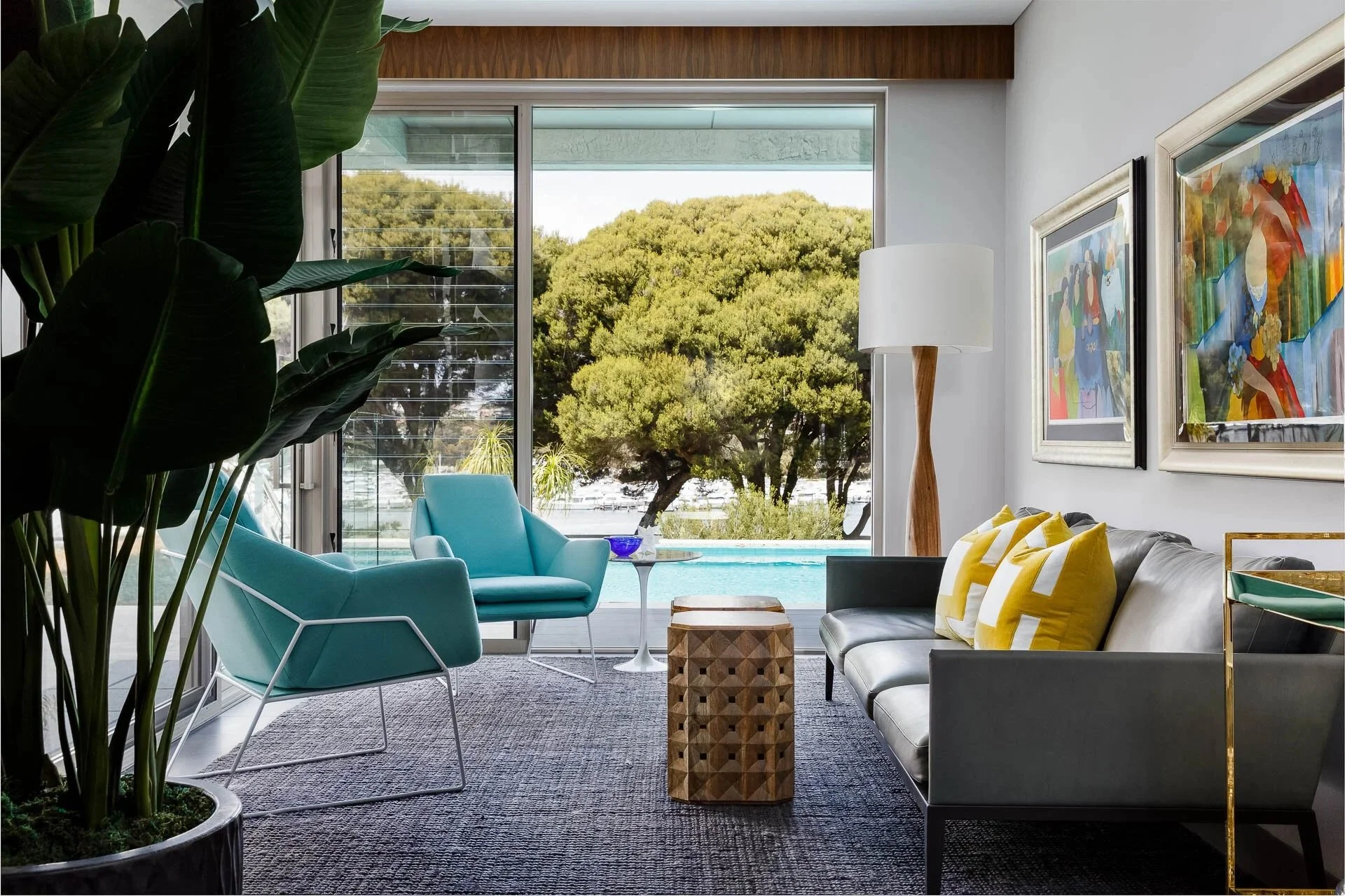 Poolside living room at Mathieson Residence featuring turquoise lounge chairs, a contemporary leather sofa, colourful artwork and seamless indoor-outdoor connections overlooking landscaped gardens and a private swimming pool.