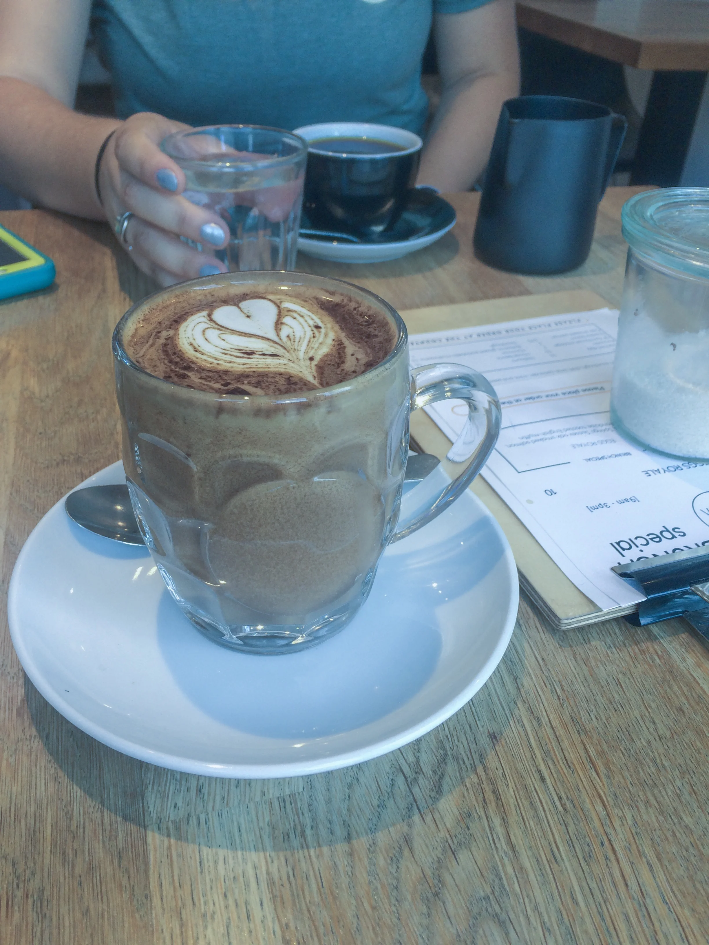 Image Description: a mocha in a glass cup with a heart design on the top sits on the table closest to the camera. Across the table, there is someone in a blue t-shirt with a cup of coffee in a black cup in front of them. Only their chest is visible. They hold a water glass in their left hand, nails painted with silver polish. A menu sits on the table next to them, with a glass jar full of sugar on top of it.