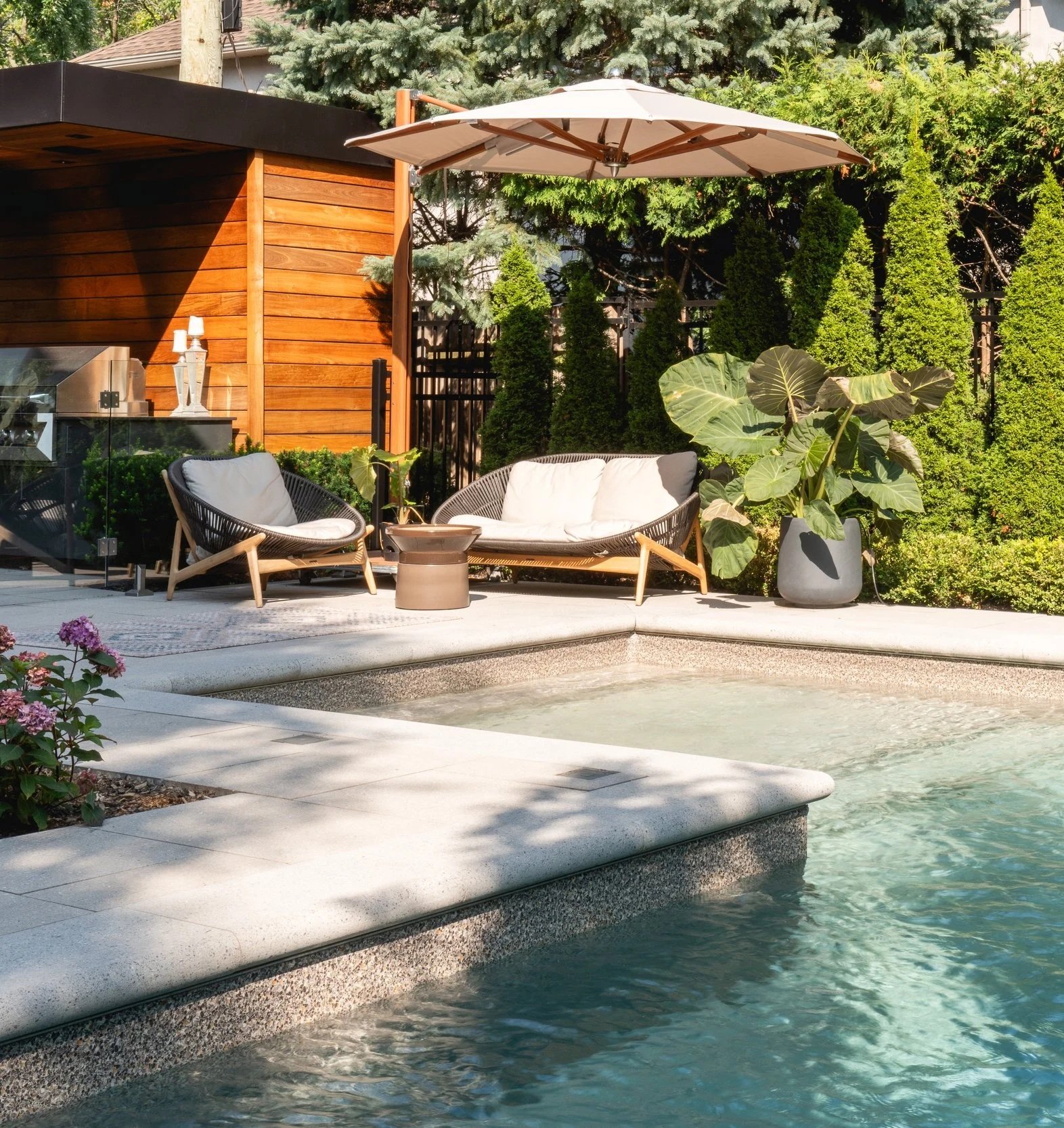 Coin salon extérieur avec des meubles en rotin, un parasol, des plantes vertes, une piscine et un mur en bois.