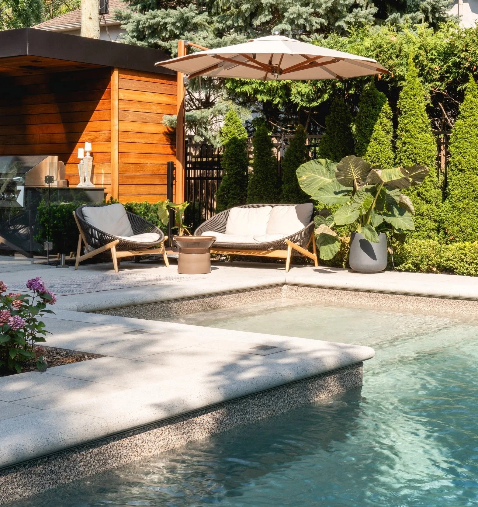 Coin salon extérieur avec des meubles en rotin, un parasol, des plantes vertes, une piscine et un mur en bois.