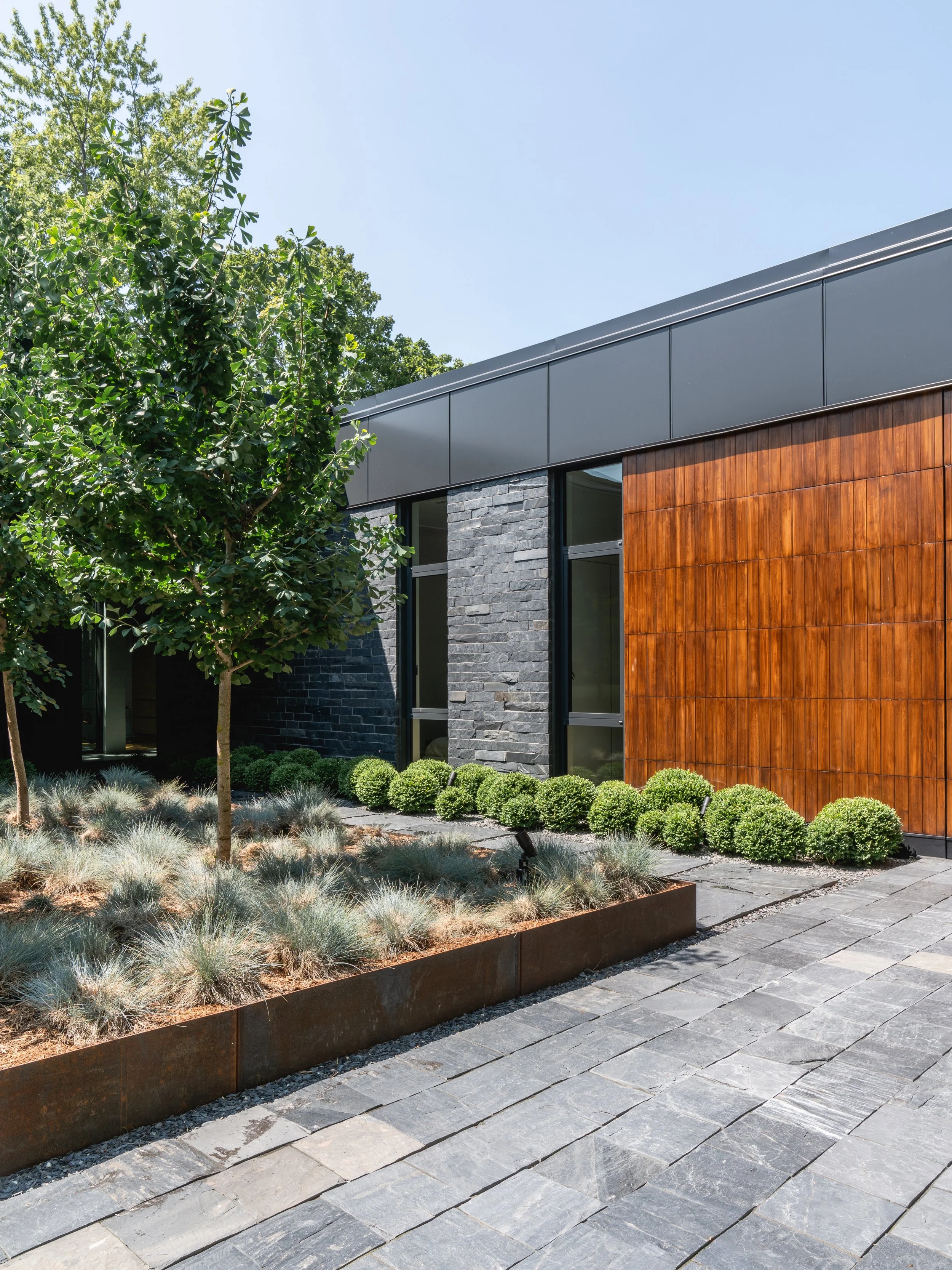 Façade moderne d'un bâtiment avec mélange de pierre noire, bois chaud et panneaux métalliques gris, entouré de plantes ornementales, y compris des arbres et des arbustes verts, sous un ciel clair.