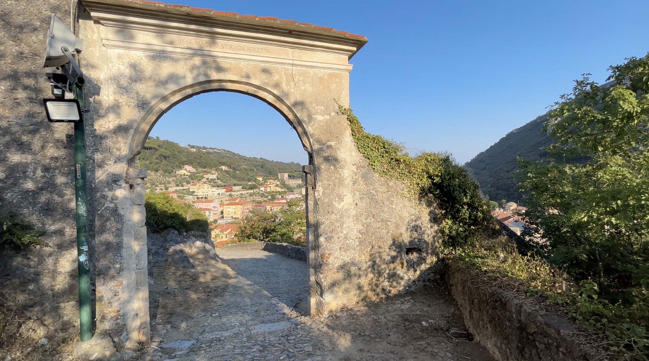 Stone archway framing colorful buildings and wooded hills in Finale Ligure, Italy, with a cobbled path passing through.