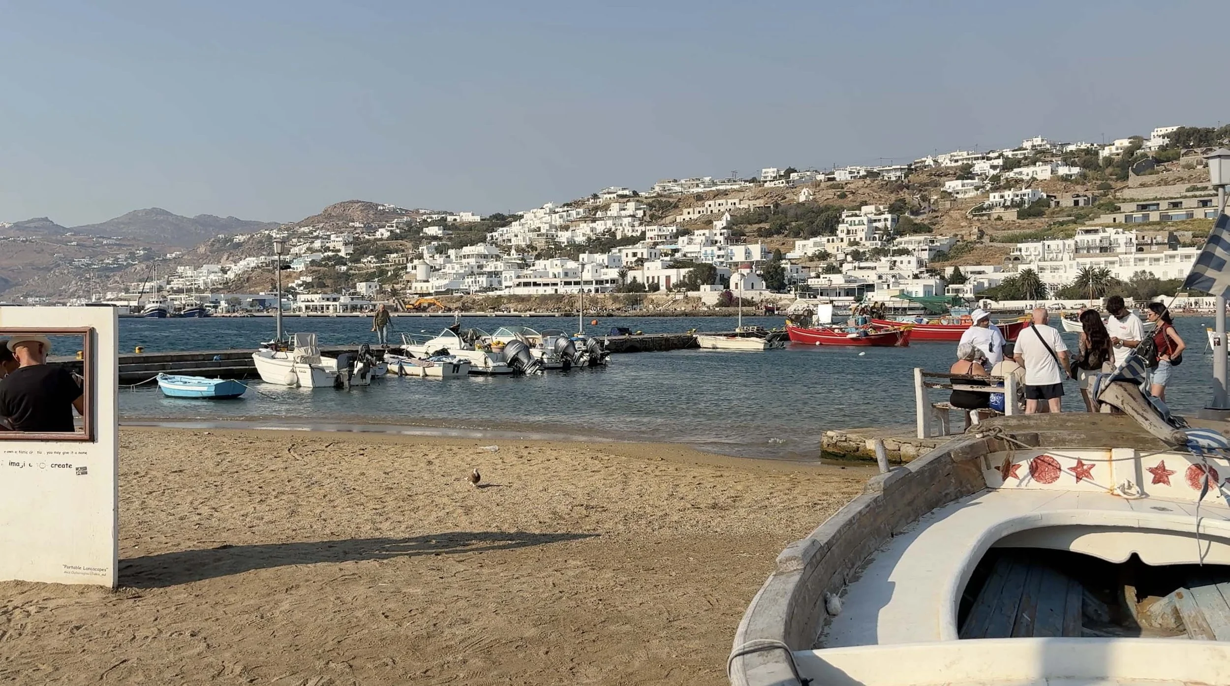 Small boats beside a sandy waterfront in Mykonos, Greece, with white buildings across the harbor and surrounding hills.